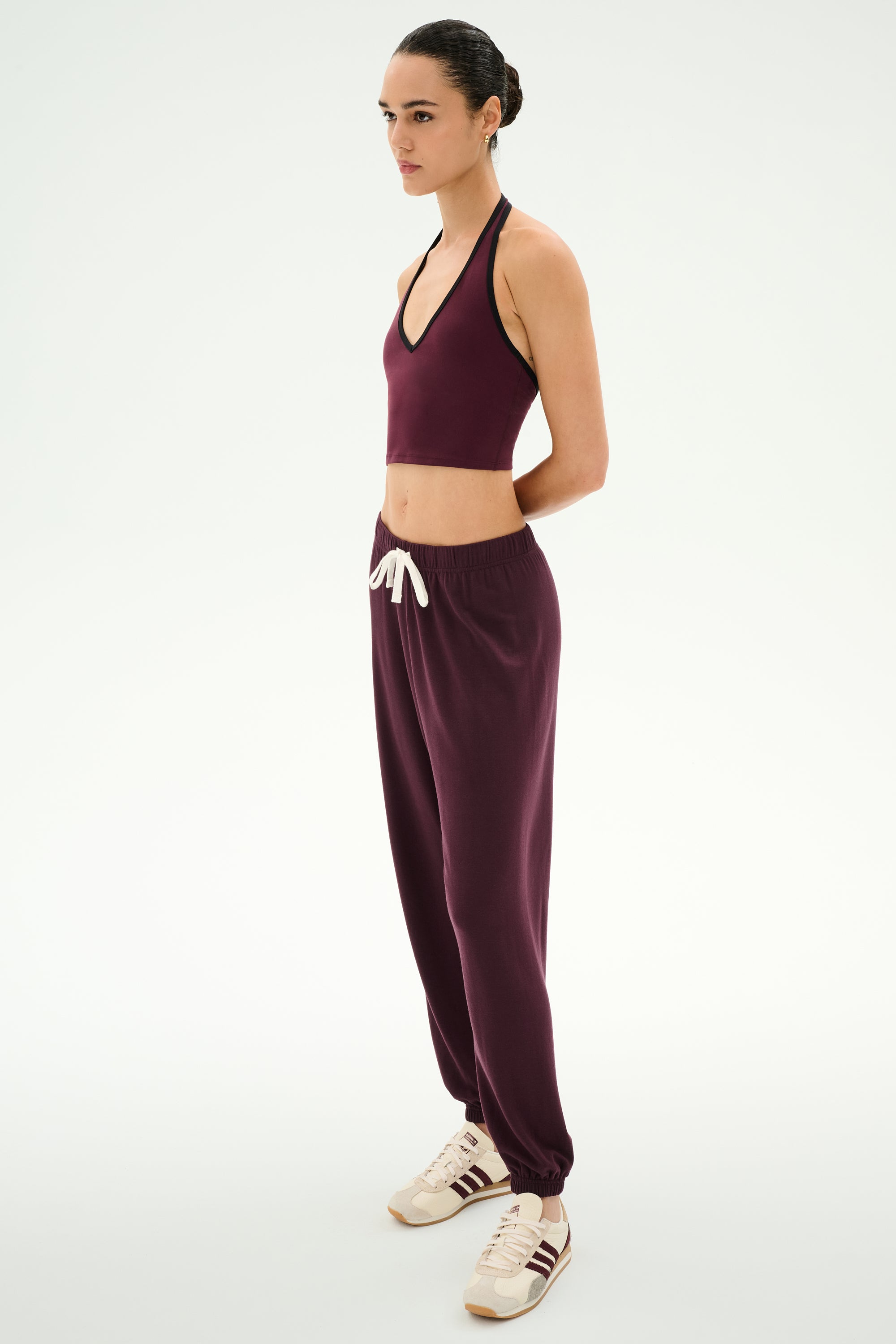 A person stands in profile wearing a maroon halter crop top, the SPLITS59 Andie Oversized Fleece Sweatpant in Dark Cherry with a white drawstring, and white sneakers with maroon stripes—perfect for cool weather workouts.