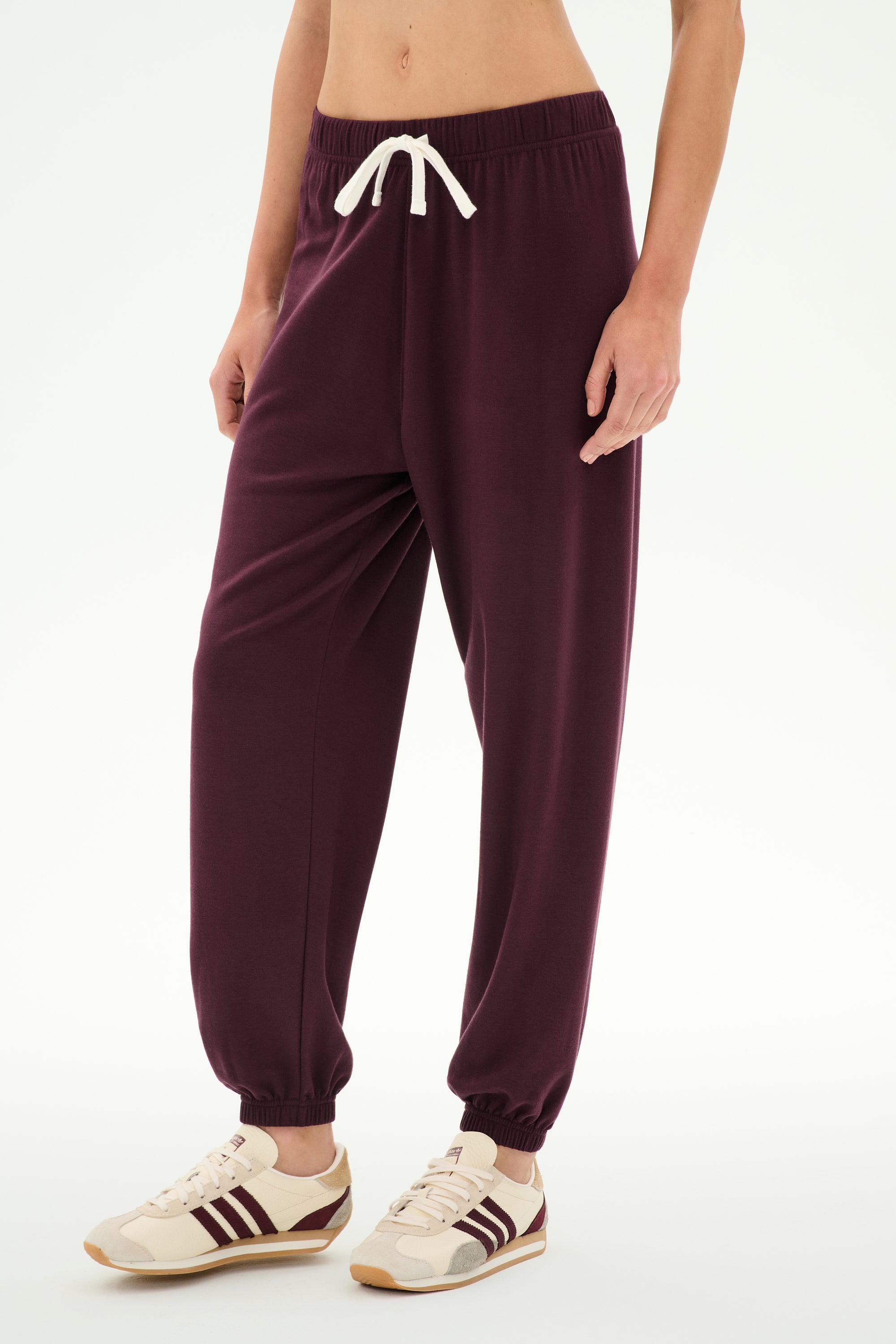 Wearing the SPLITS59 Andie Oversized Fleece Sweatpant in Dark Cherry, featuring a white drawstring, this outfit is paired with cream sneakers accented by brown stripes. The ultra-luxe fleece joggers, crafted in Los Angeles, are ideal for cool weather workouts and are shown against a plain white background.