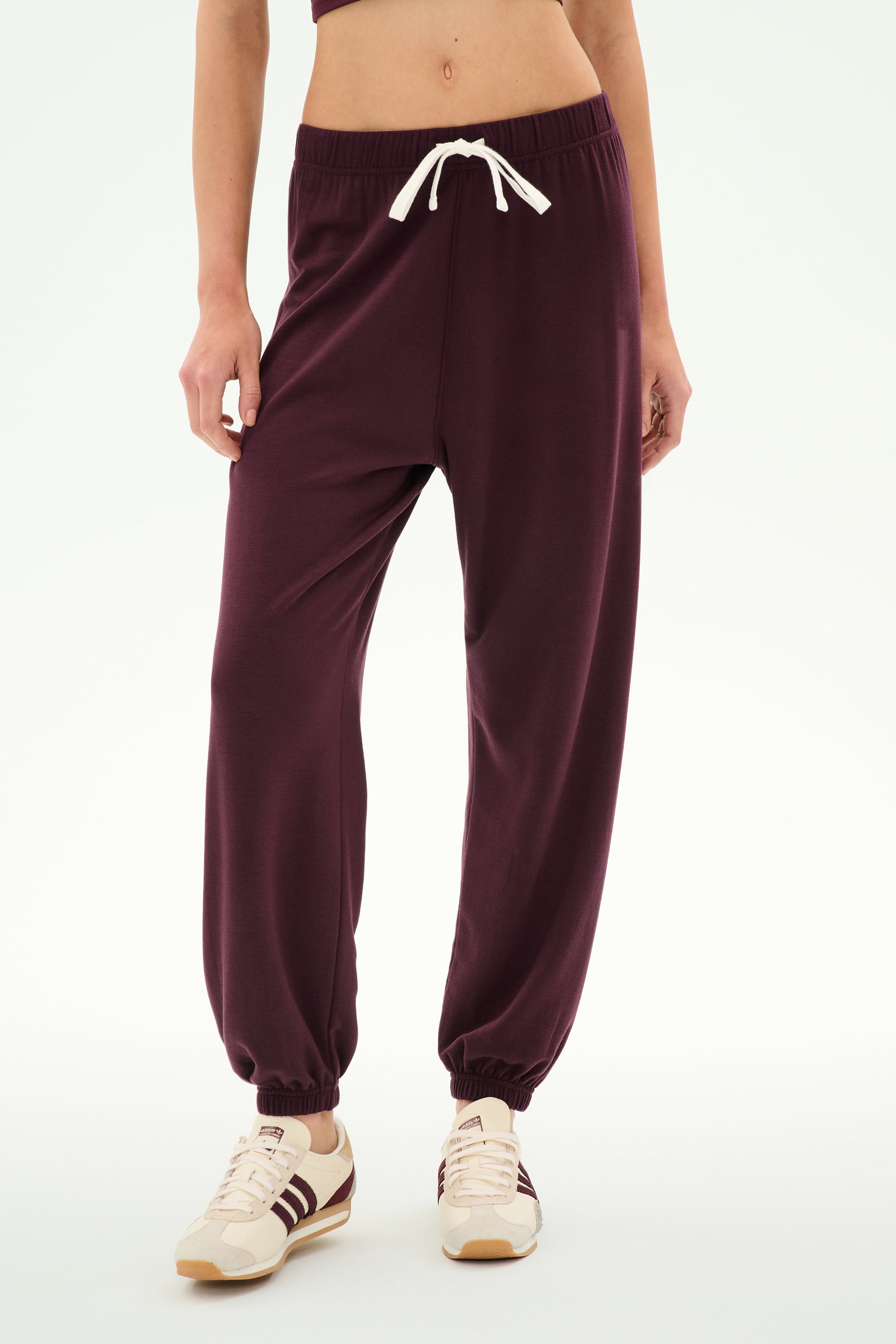 A person stands against a white background wearing SPLITS59′s Andie Oversized Fleece Sweatpant in Dark Cherry—crafted from ultra-luxe fleece and made in Los Angeles—paired with beige sneakers featuring burgundy stripes and a white drawstring; ideal for cool weather workouts.