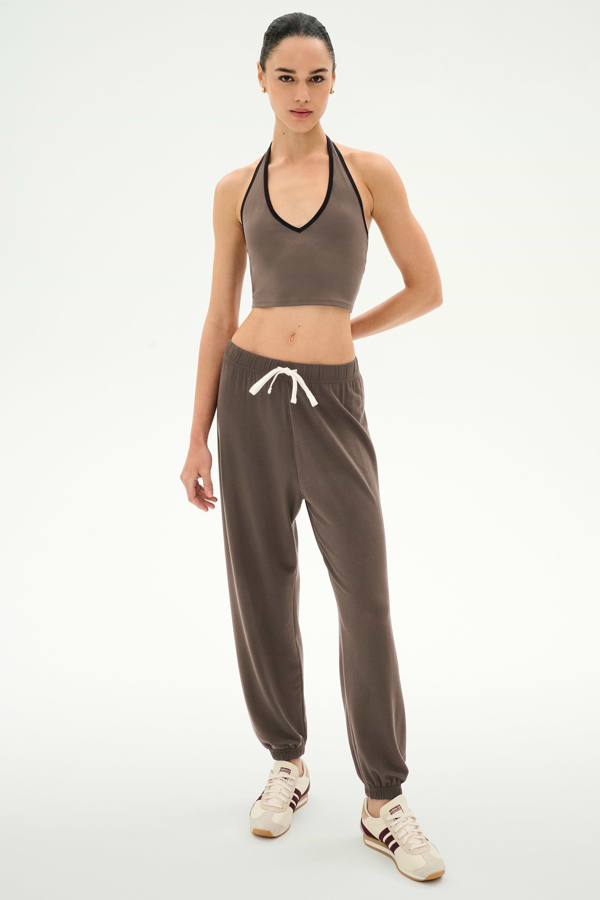 A woman stands facing forward against a plain white background, wearing SPLITS59 Andie Oversized Fleece Sweatpant in Lentil with a brown halter crop top and white sneakers.