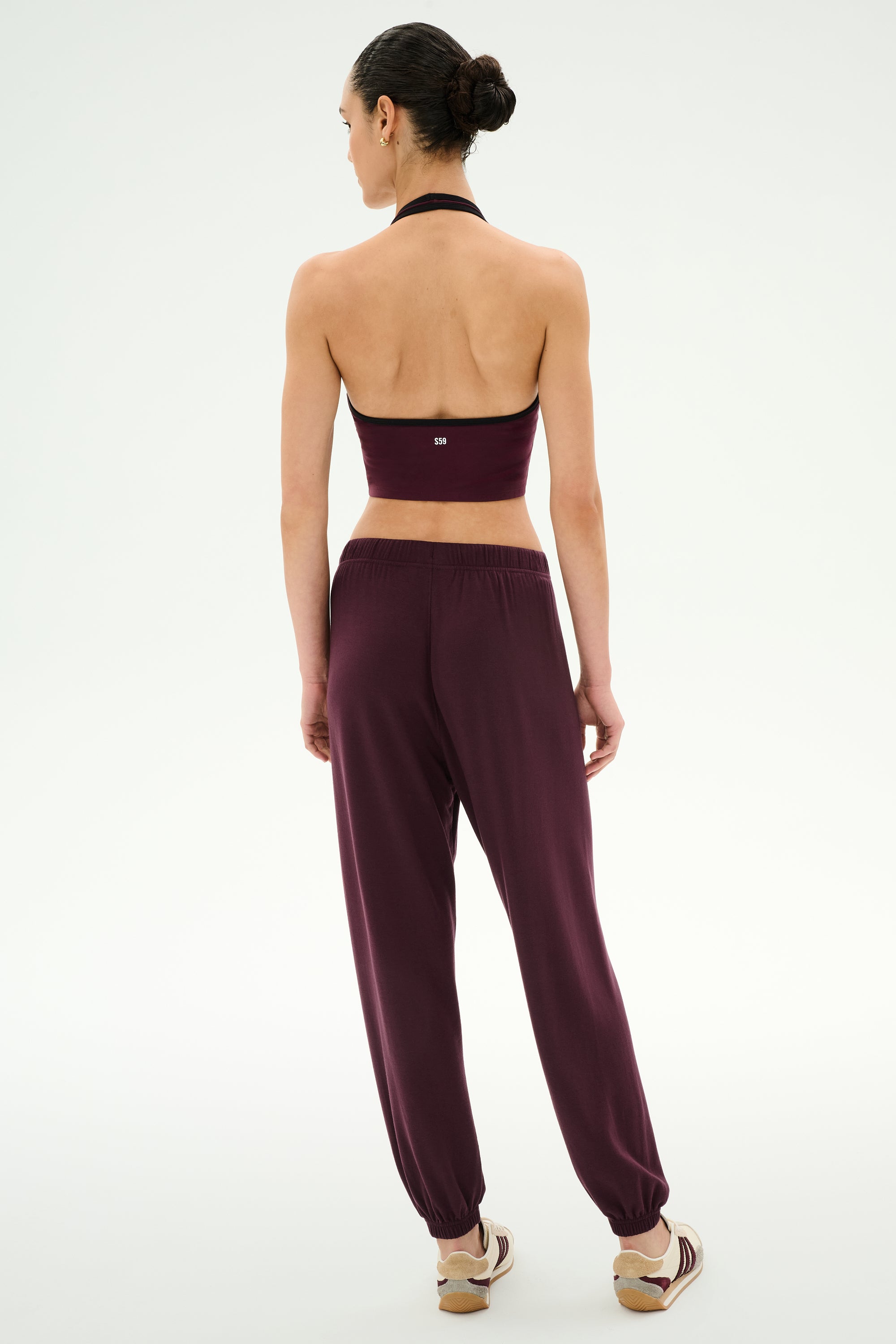 A woman stands facing away, wearing a dark purple halter top and SPLITS59′s Andie Oversized Fleece Sweatpant in Dark Cherry with elastic ankles—perfect for cool weather workouts—paired with beige flats.