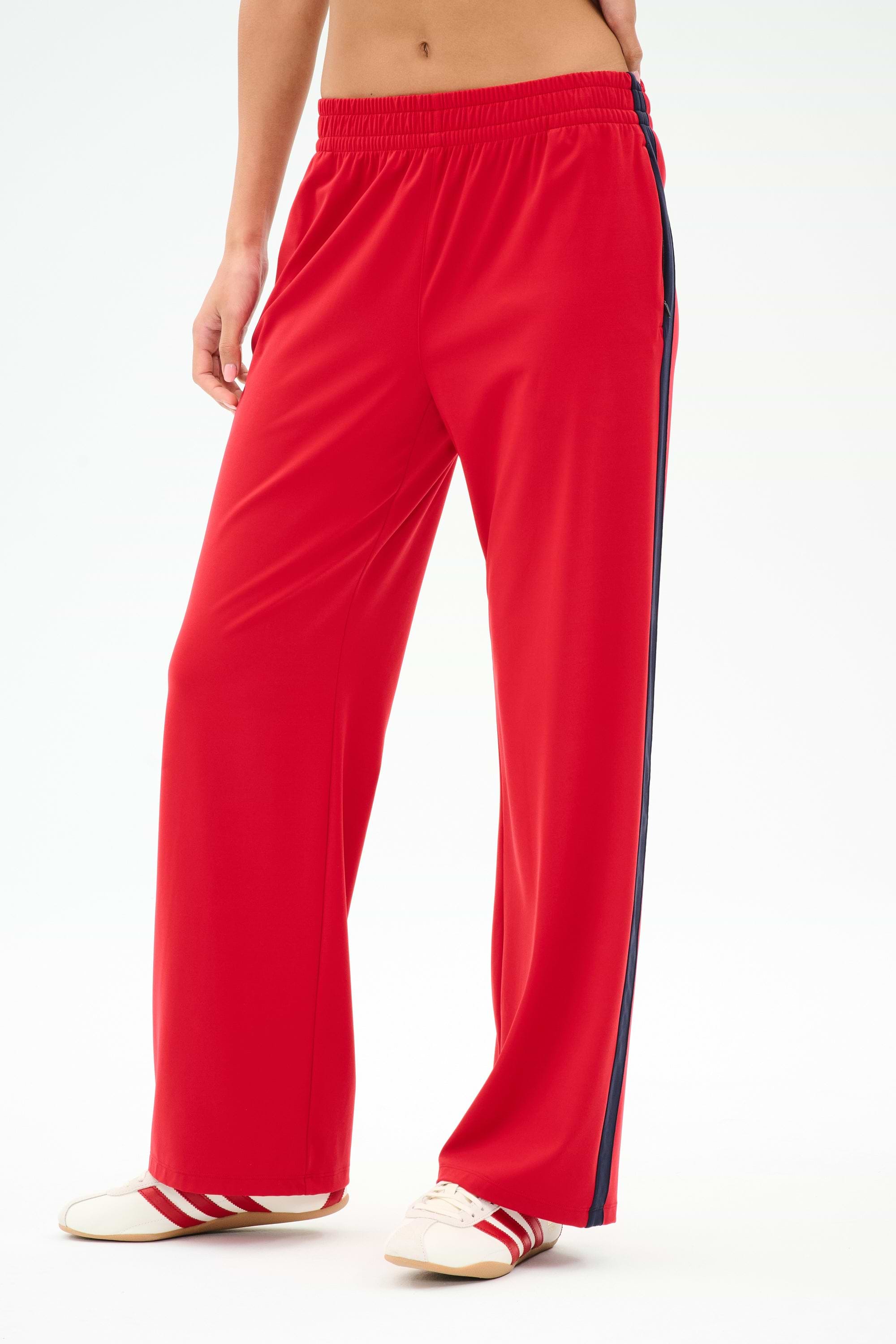 A person wearing SPLITS59 Maxie Rigor Track Pant in Pirate Red/Indigo, paired with white sneakers featuring red accents, stands against a plain background.