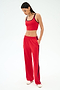 A woman stands against a white background wearing a red sports bra, SPLITS59 Maxie Rigor Track Pant in Pirate Red/Indigo with side stripes, and white sneakers, hands in her pockets and looking down.