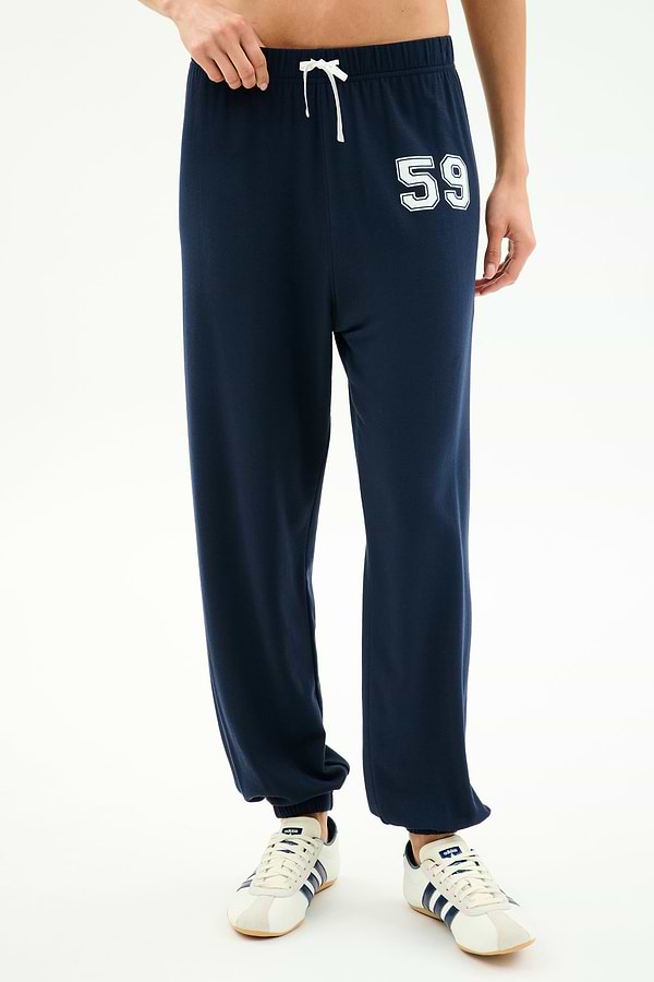 Wearing SPLITS59 Andie Oversized Fleece Varsity 59 Sweatpant in Indigo with a white drawstring and "59" on the left thigh, paired with white sneakers featuring navy stripes. Made in Los Angeles for superior comfort and style.