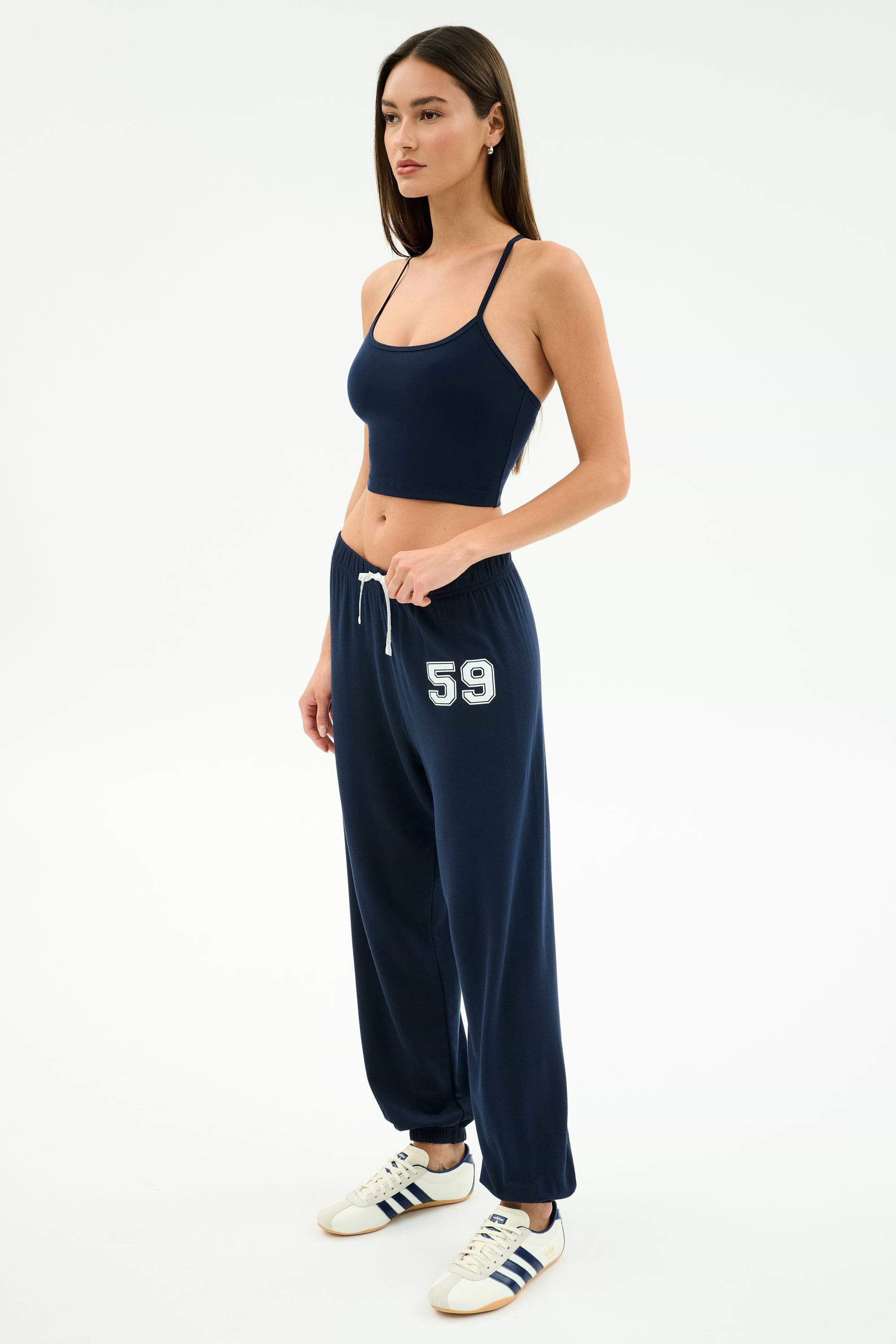 A woman wears the SPLITS59 Andie Oversized Fleece Varsity 59 Sweatpant in Indigo/59, paired with a navy crop top and white sneakers with navy stripes, standing against a plain white background. Made in Los Angeles.
