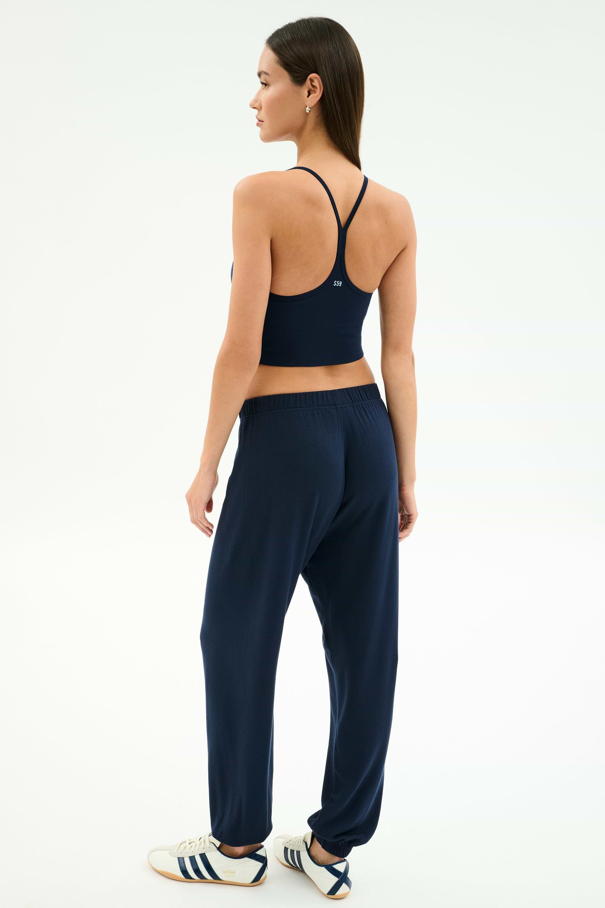 A woman faces away, wearing a navy sports bra, SPLITS59 Andie Oversized Fleece Varsity 59 Sweatpant in Indigo/59 made in Los Angeles, and white Adidas sneakers against a plain white background.
