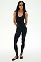 A woman with long braids faces forward, wearing the SPLITS59 Darci Airweight Stirrup Jumpsuit in black and black ballet flats, on a plain white background.