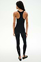 A woman faces away in the SPLITS59 Darci Airweight Stirrup Jumpsuit - Black, a fitted, stretch black sleeveless jumpsuit paired with black ballet flats, set against a plain white background.