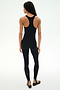 A woman faces away in the SPLITS59 Darci Airweight Stirrup Jumpsuit - Black, a fitted, stretch black sleeveless jumpsuit paired with black ballet flats, set against a plain white background.