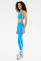 A woman stands in profile against a white background wearing the SPLITS59 Ella Airweight Bra in Riviera/White with matching high-waisted leggings, white socks, and gray sneakers—an ideal look for hot yoga.