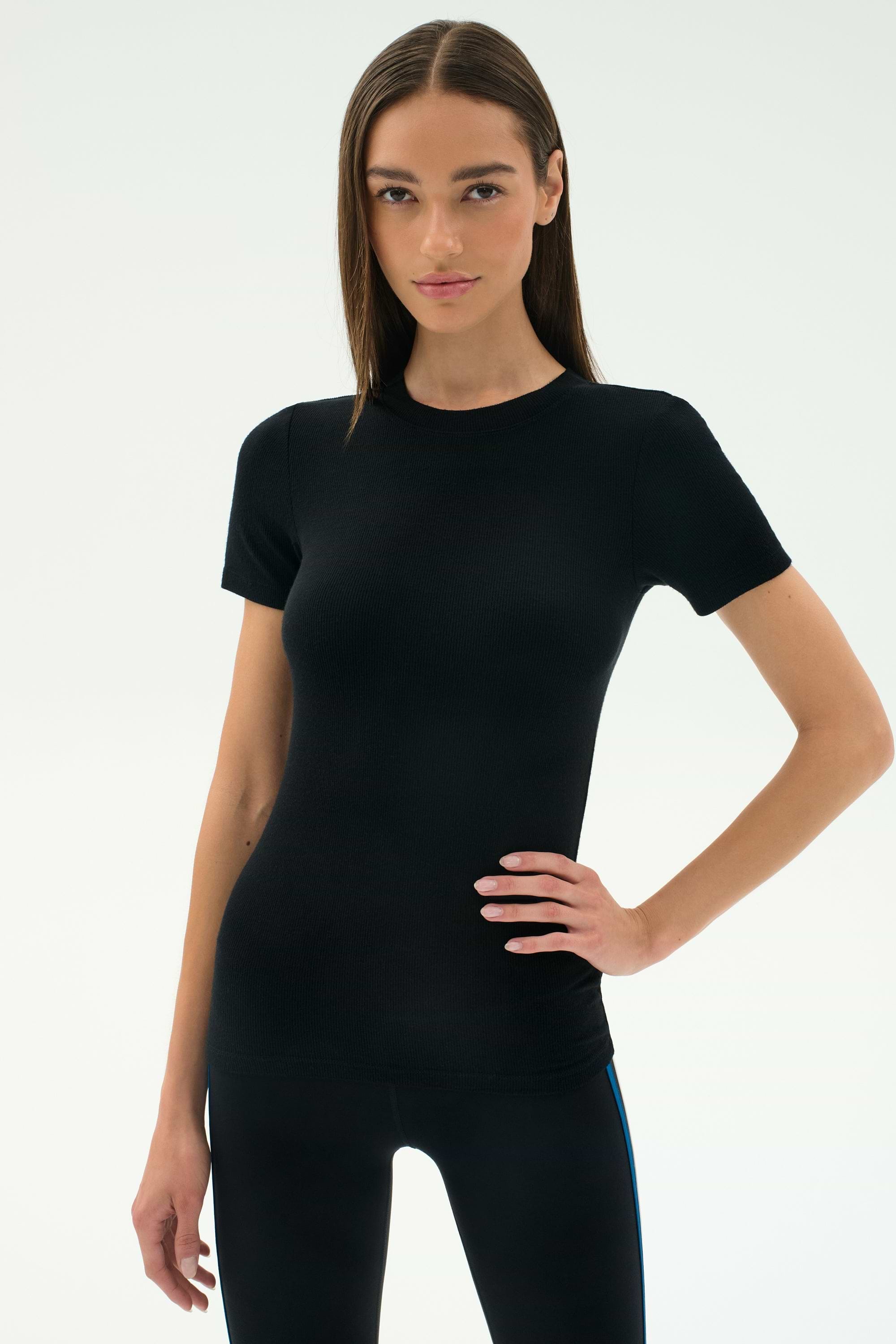 A woman poses against a plain background wearing the SPLITS59 Louise Rib Short Sleeve in black with matching pants, one hand on her hip.