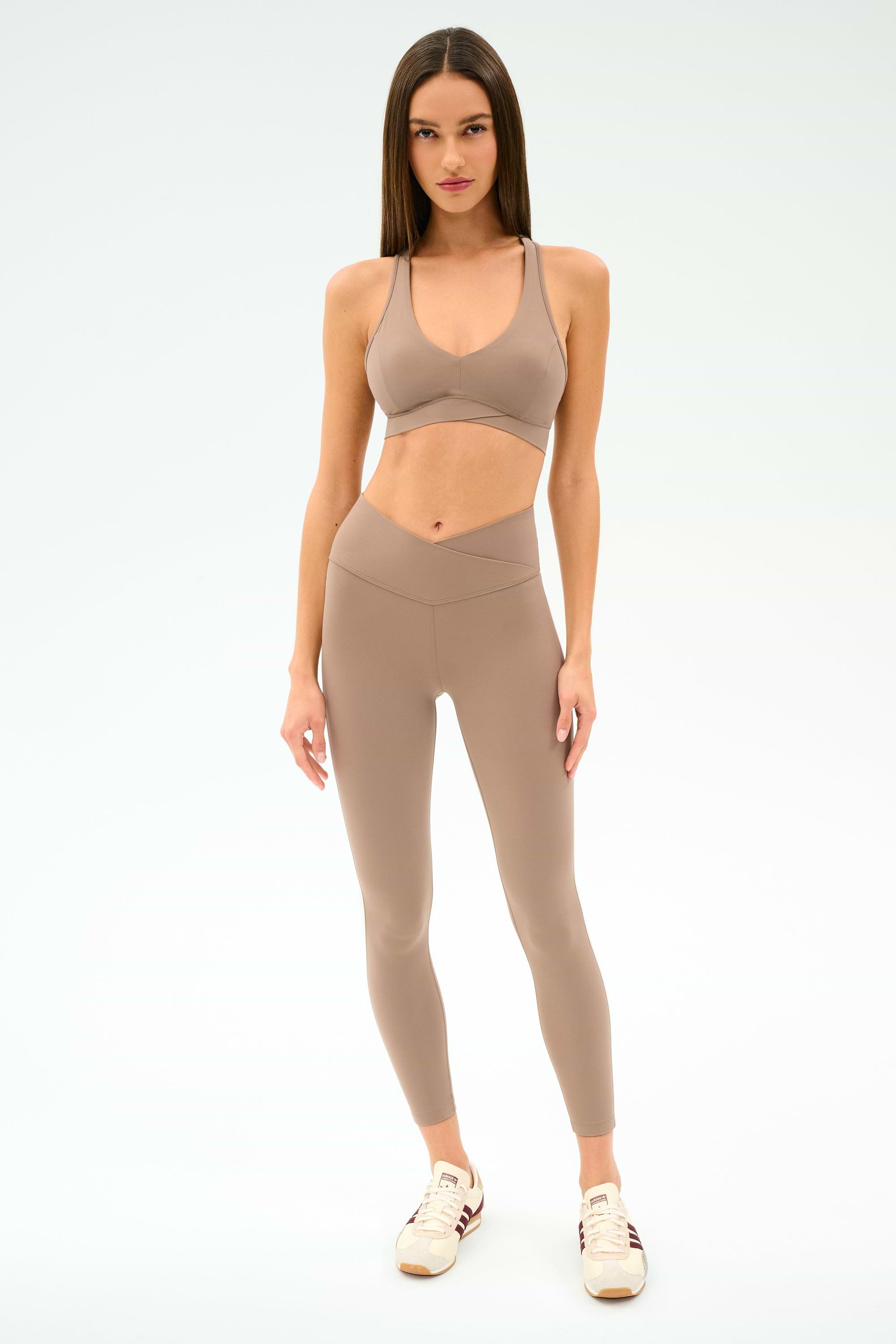 A woman faces the camera wearing the SPLITS59 Mia Rigor Bra in Latte, with matching high-waisted leggings and beige sneakers against a plain light background.
