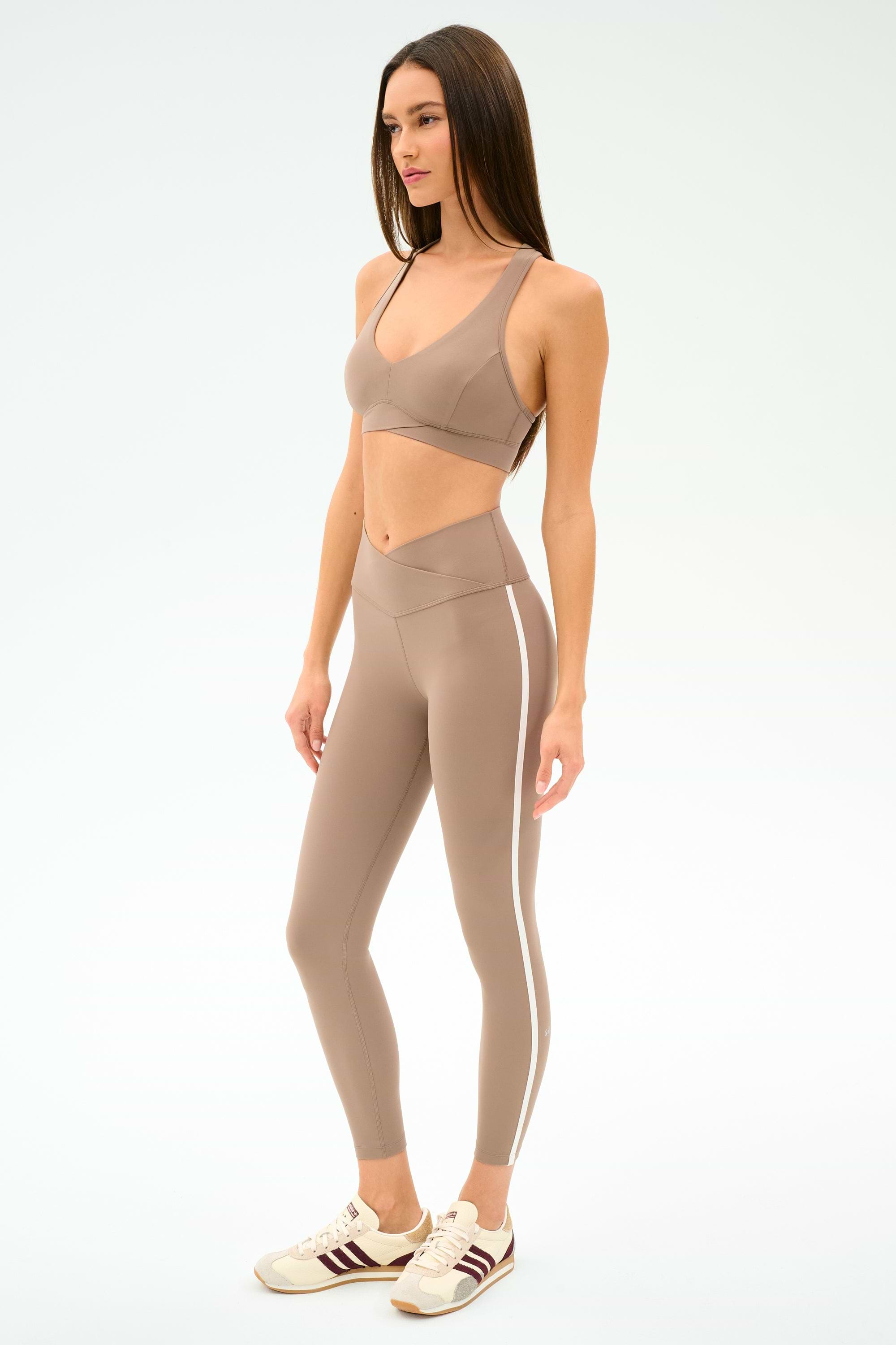 A woman wears SPLITS59's Mia Rigor Legging w/ Stripe in Latte/White with a taupe sports bra and white sneakers with brown accents, standing against a plain white background.