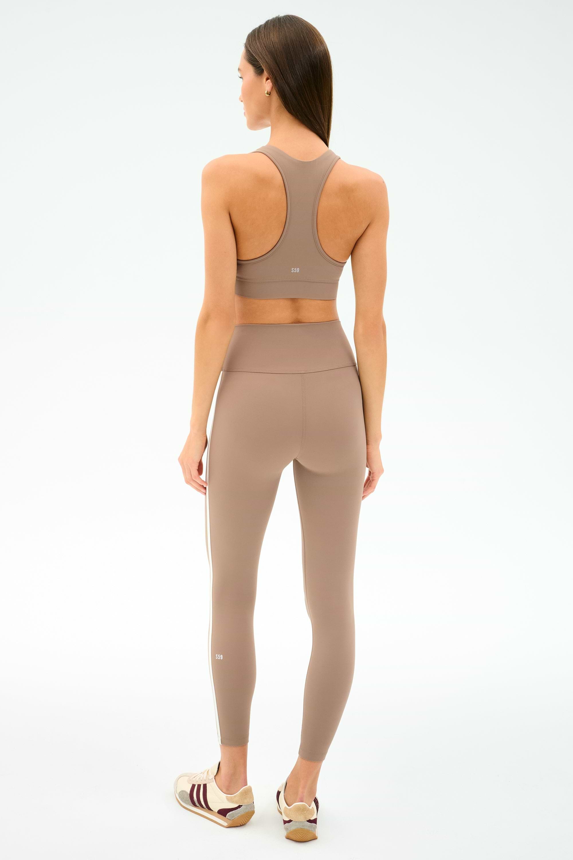 A woman faces away, wearing the SPLITS59 Mia Rigor Bra in Latte with matching leggings and white athletic shoes, set against a plain light background.