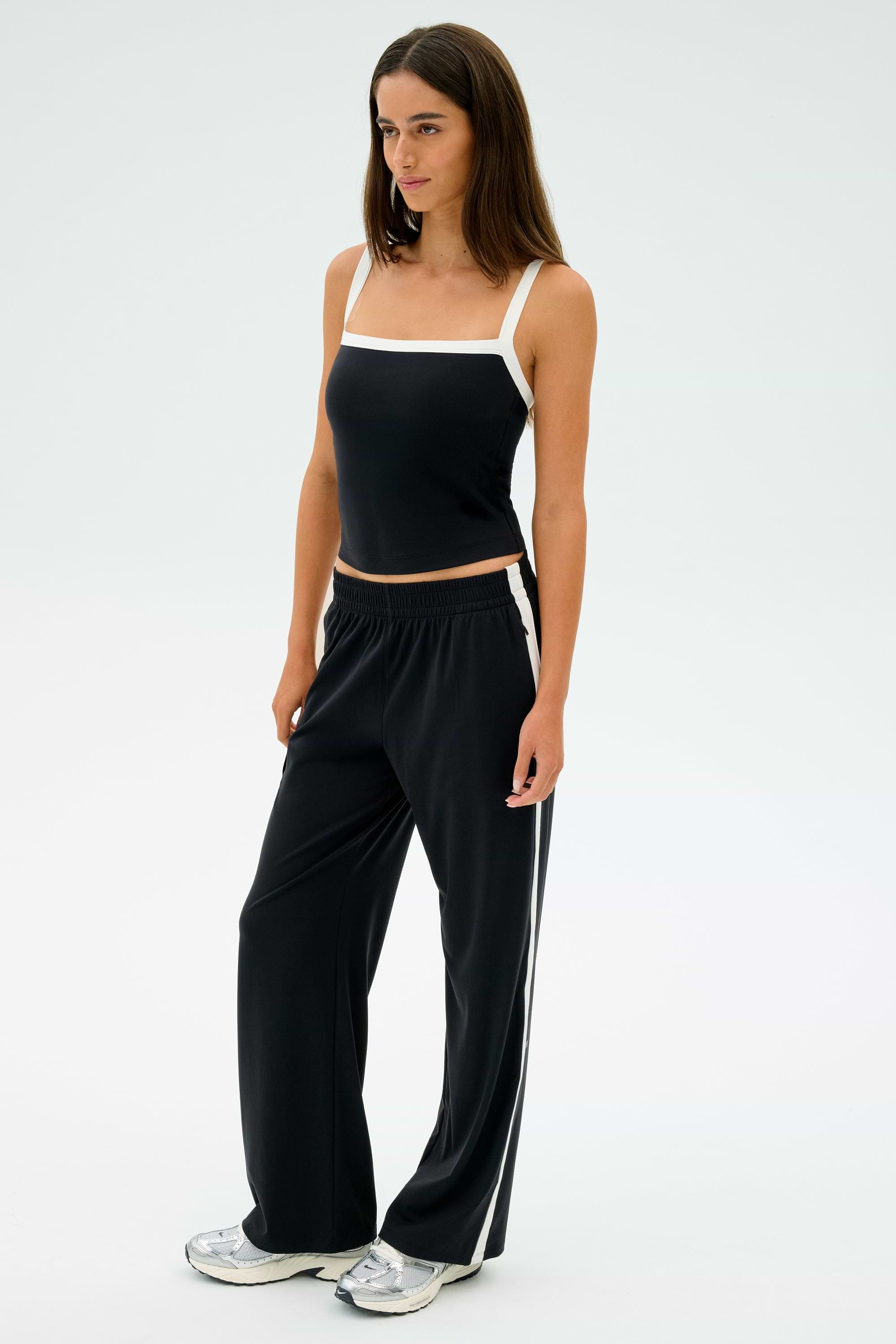 A person stands wearing a sleeveless black top, SPLITS59 Maxie Rigor Track Pants in Black/White, and silver sneakers against a plain white background.