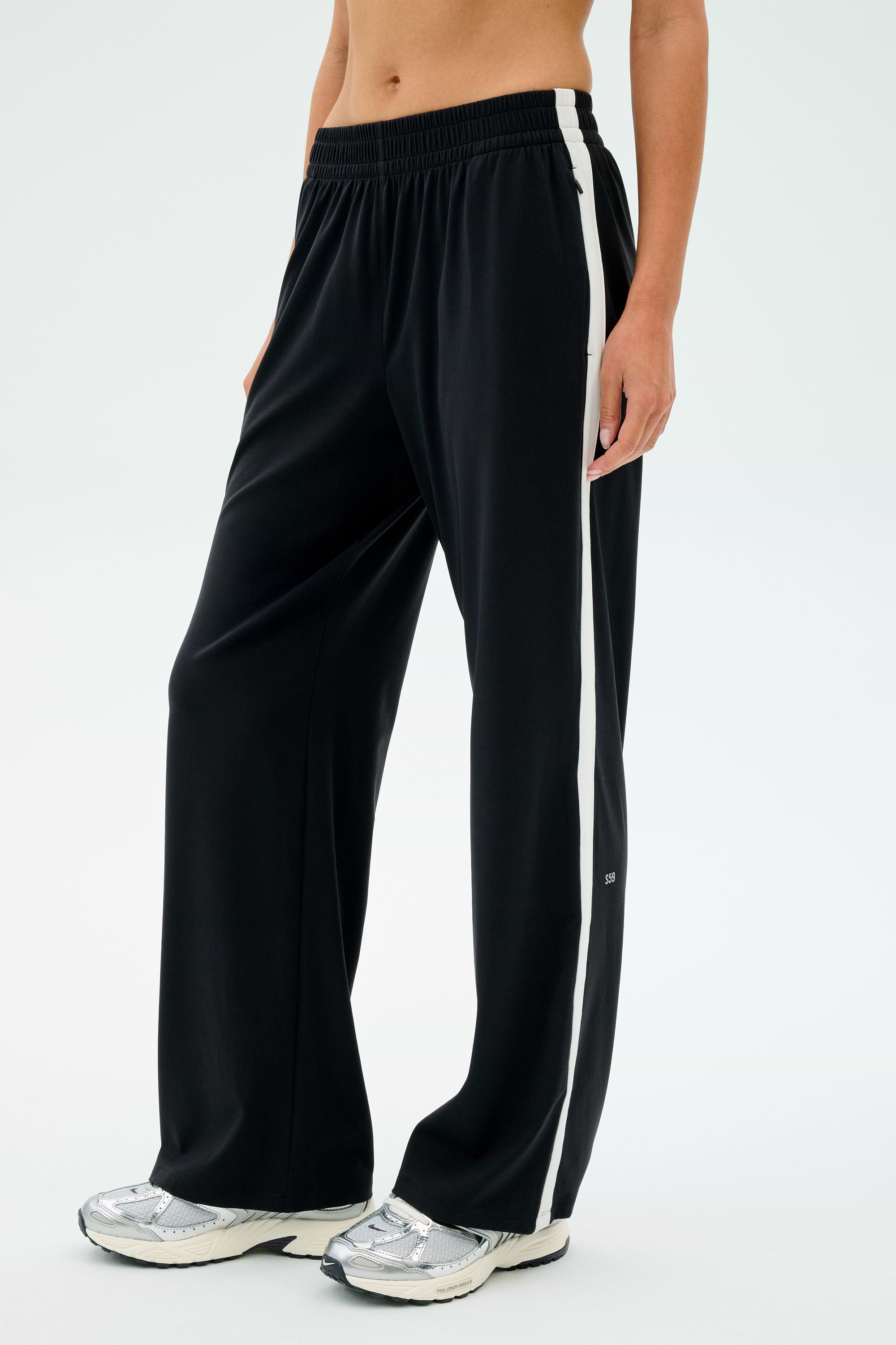 A person wearing the SPLITS59 Maxie Rigor Track Pant in Black/White with silver sneakers stands against a plain background.