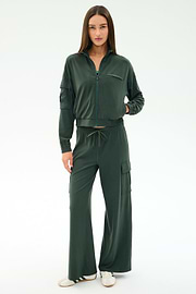 A woman faces forward in SPLITS59’s Alex Airweight Cargo Pant - Military, styled with a dark green nylon zip-up jacket and white sneakers, against a plain white background.