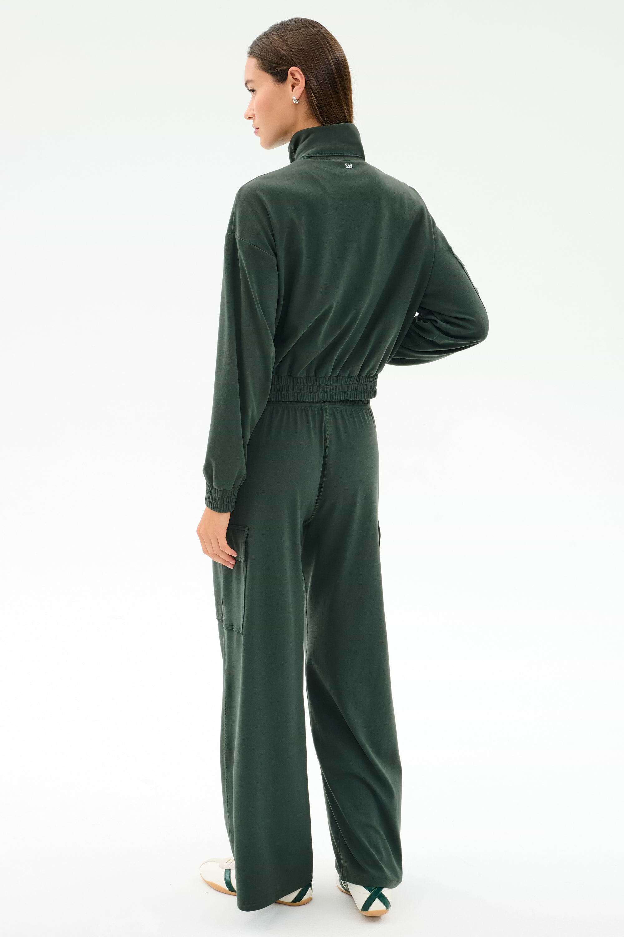 A woman stands with her back to the camera, wearing the SPLITS59 Peaches Airweight Cargo Jacket in Military, featuring soft nylon spandex, a high collar, and wide-leg pants, paired with white sneakers on a plain light background.