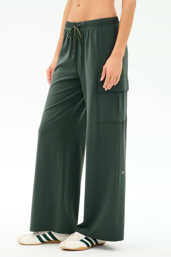 A person wears SPLITS59’s Alex Airweight Cargo Pant in Military, featuring a soft nylon blend, paired with white sneakers with green stripes, standing against a plain light background.