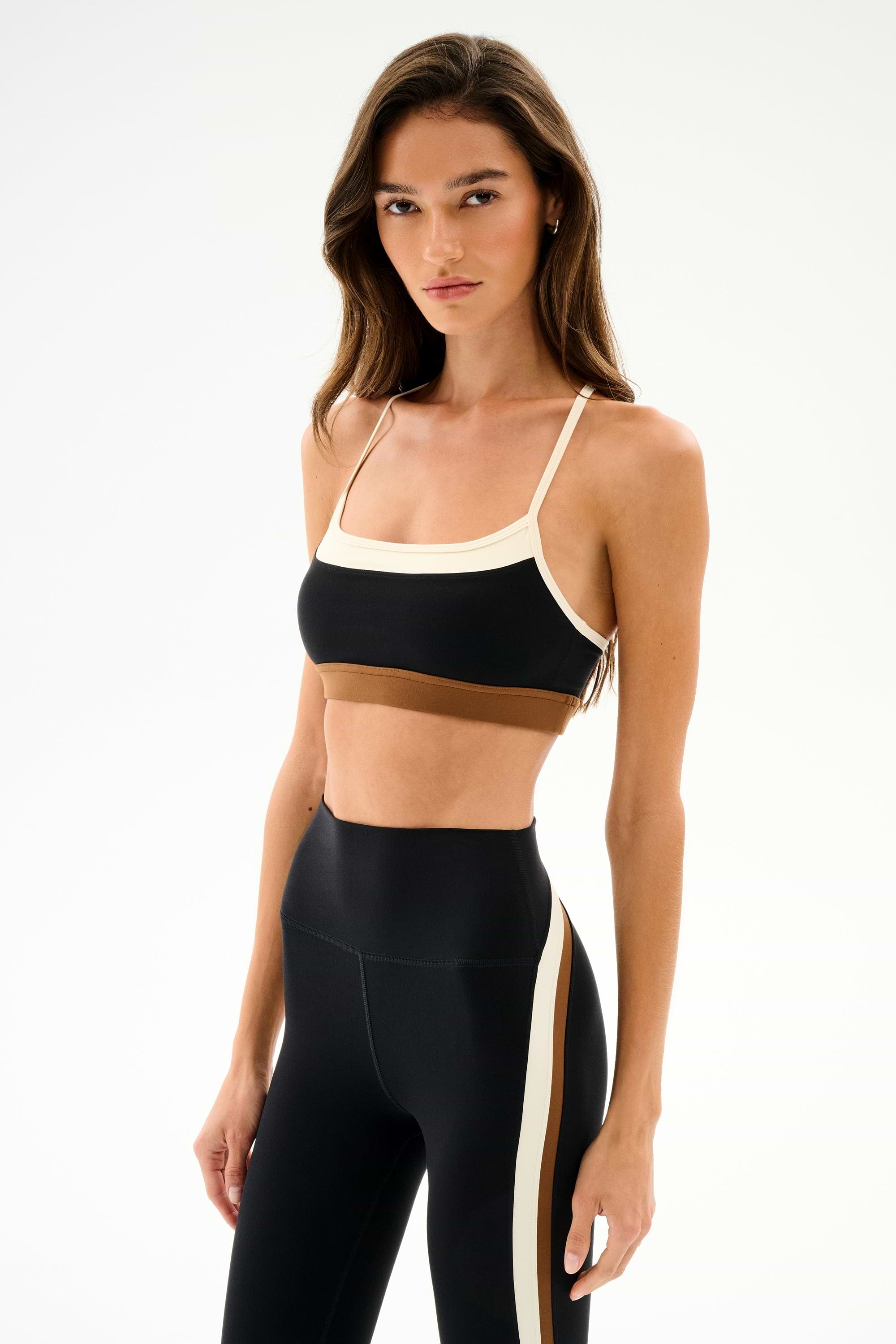 Woman in SPLITS59 Scottie Airweight Bra - Black/Creme and high-waisted black nylon leggings with a white side stripe, standing against a plain white background.