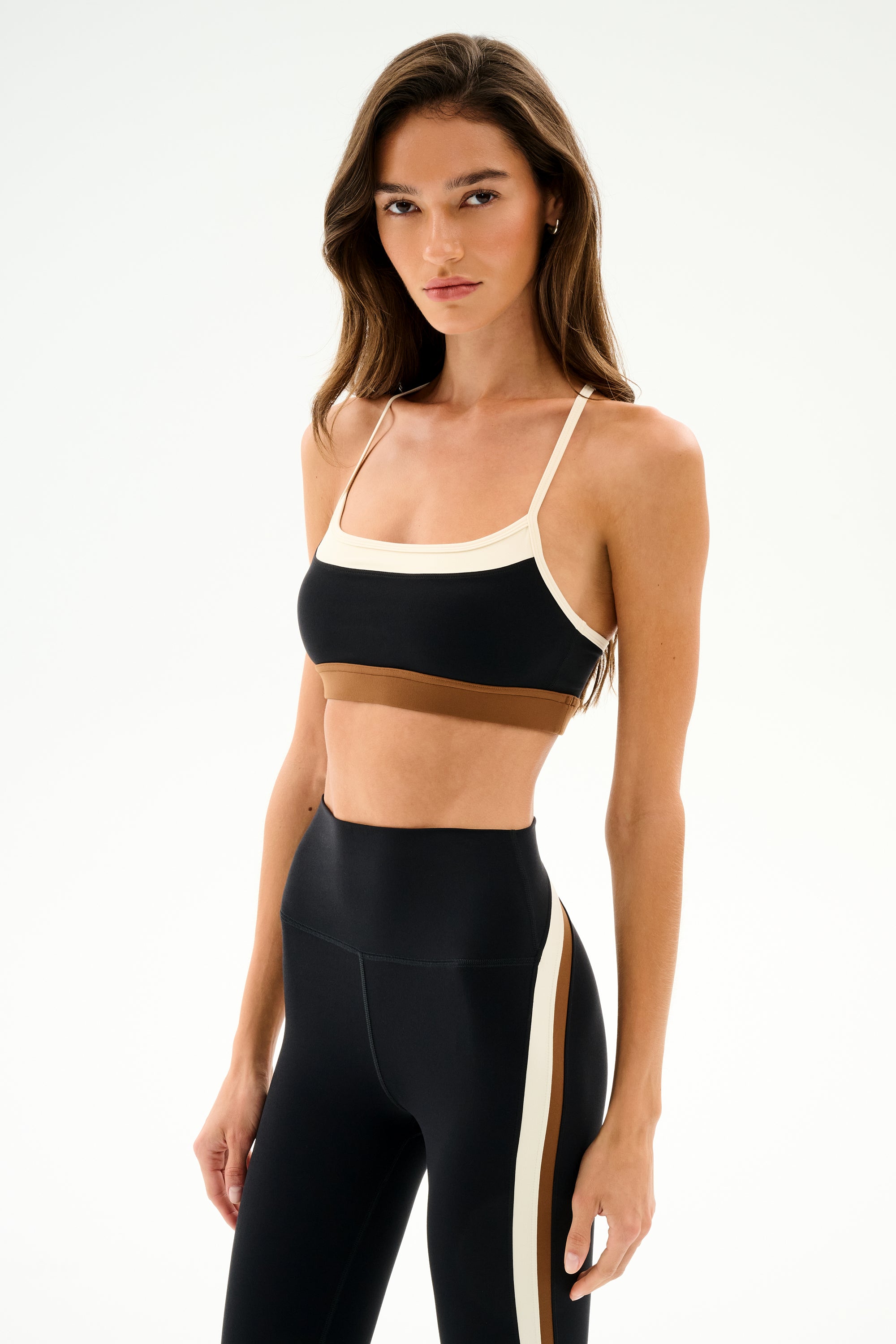 Woman in SPLITS59 Scottie Airweight Bra - Black/Creme and high-waisted black nylon leggings with a white side stripe, standing against a plain white background.