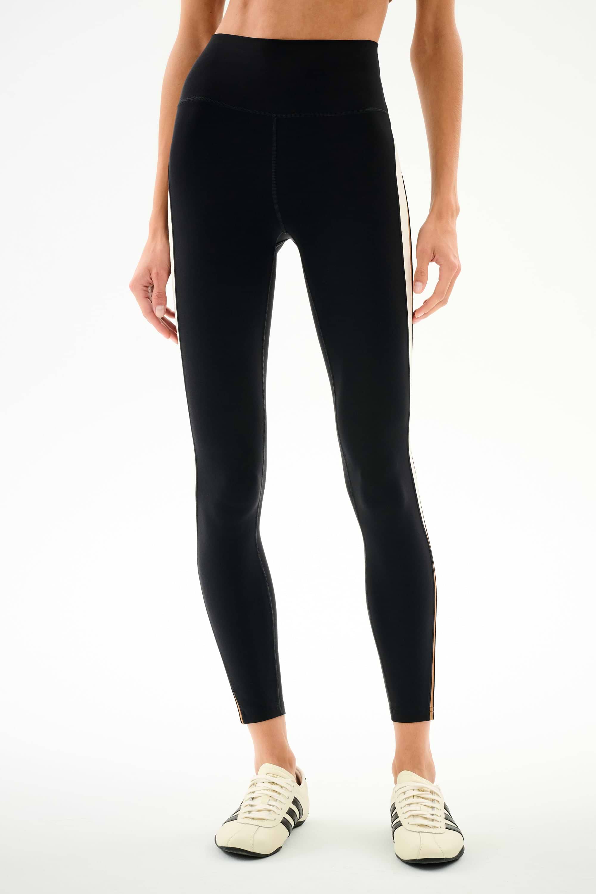 Person wearing SPLITS59 Emerson Airweight High Waist 7/8 - Black/Creme leggings with white sneakers, standing against a plain background.