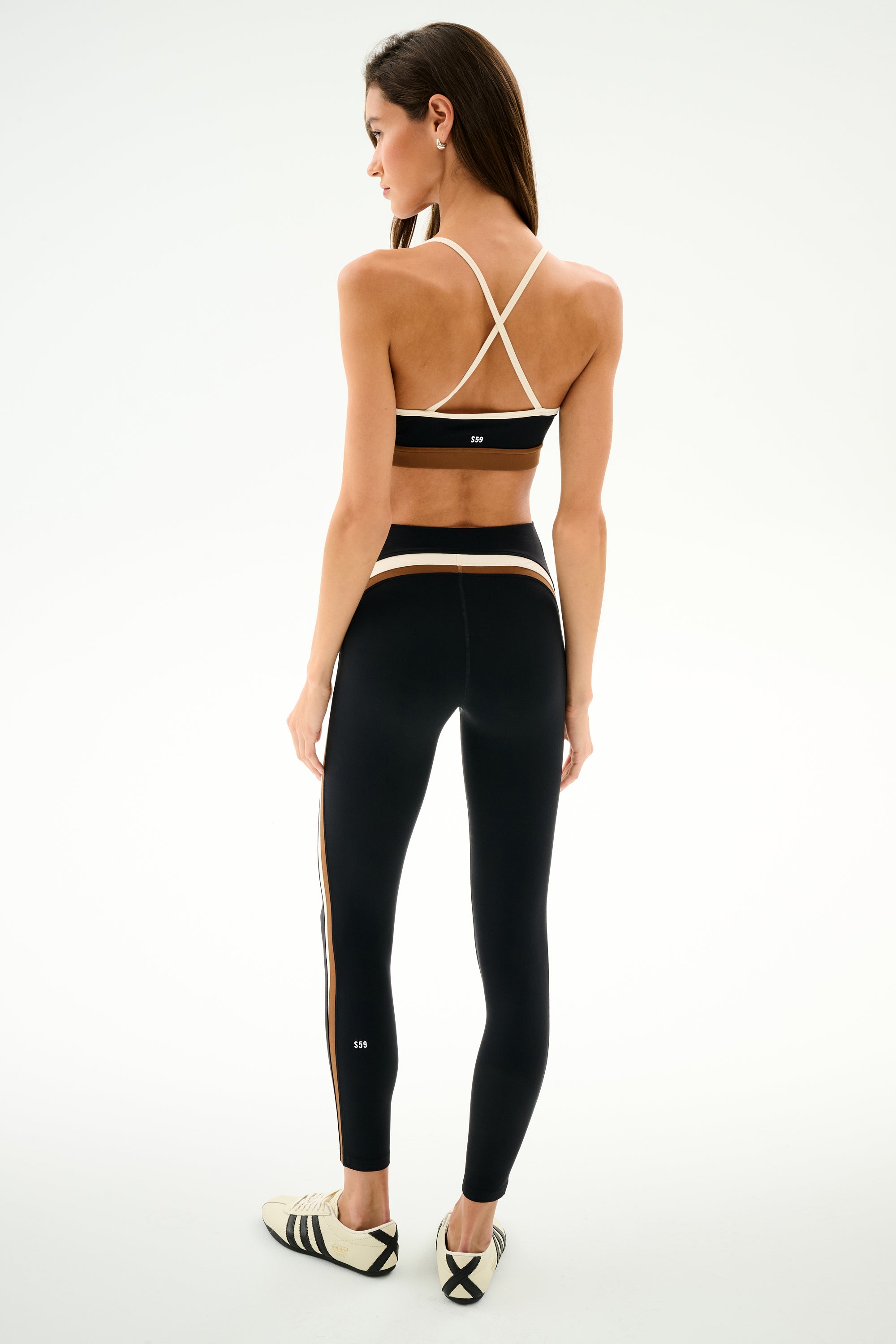 A woman faces away from the camera in SPLITS59 Emerson Airweight High Waist 7/8 - Black/Creme, styled with white sneakers against a plain white background.