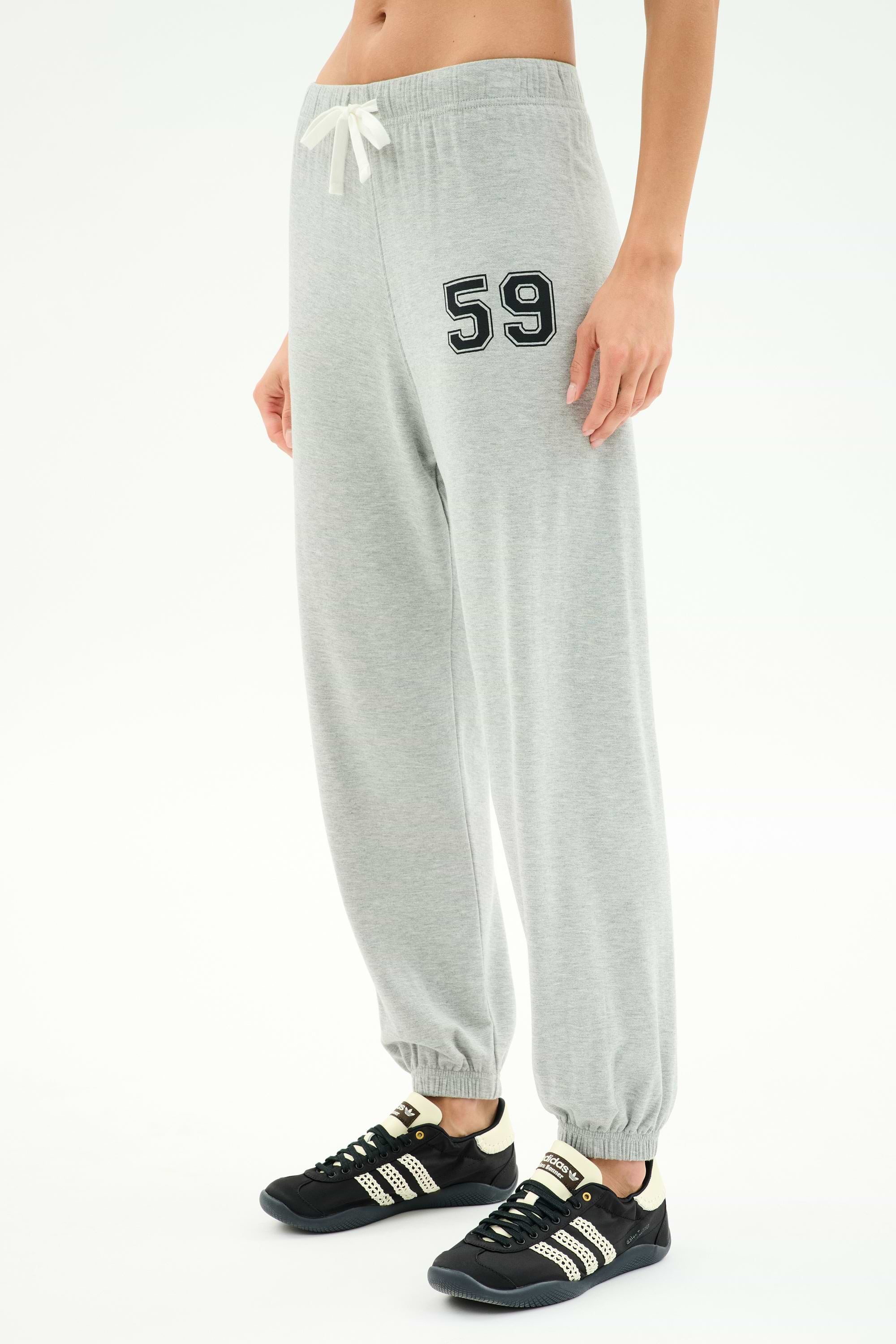 A person wearing SPLITS59 Andie Oversized Fleece Varsity 59 Sweatpant in Heather Grey/59, featuring "59" on the left thigh, white drawstring, and paired with black athletic shoes with white stripes.