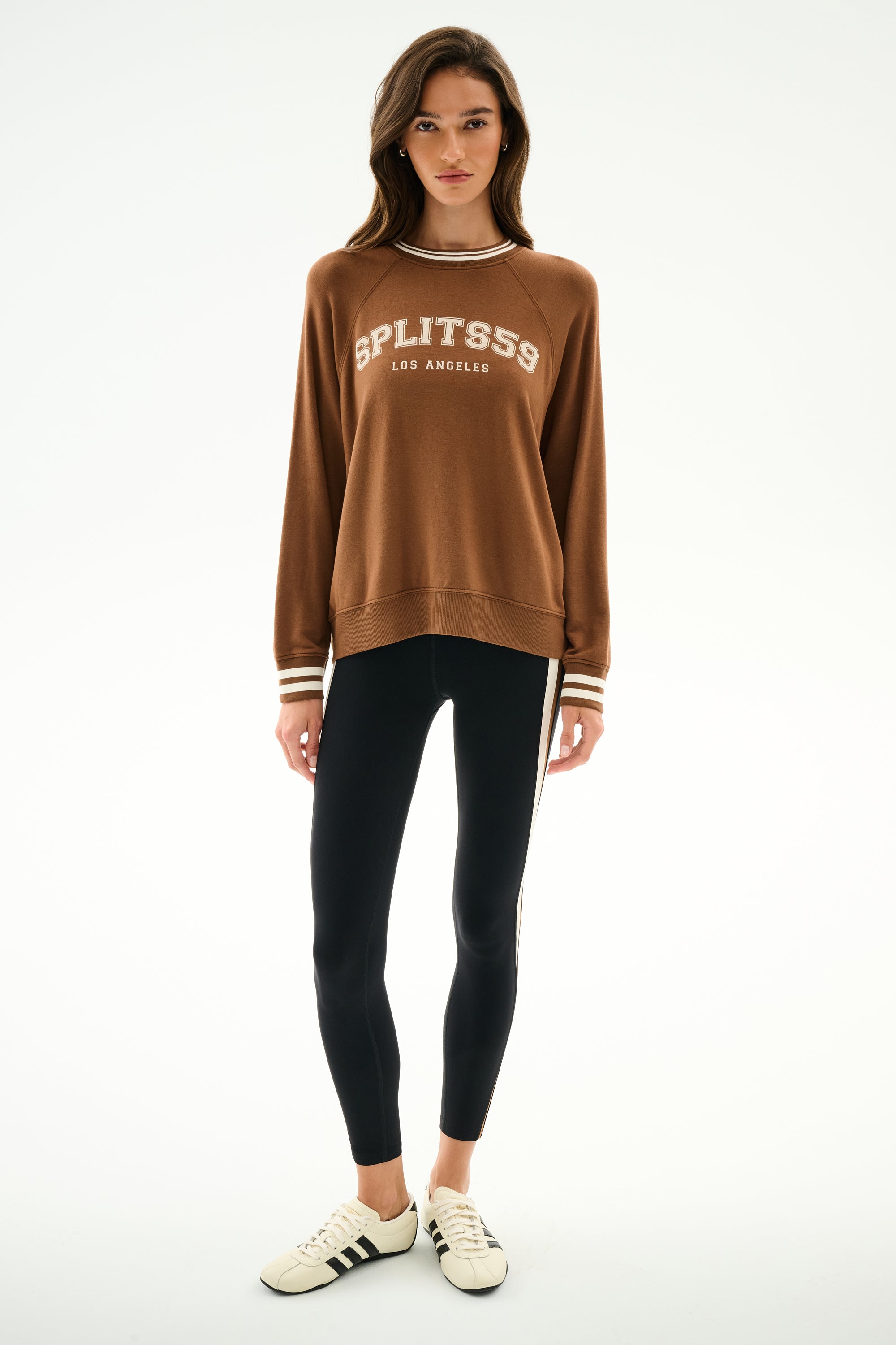 Woman stands against a white background, wearing an Andie Oversized Fleece Varsity Stripe Sweatshirt - Toffee/Splits59 Los Angeles by SPLITS59, black striped leggings, and white Adidas sneakers.
