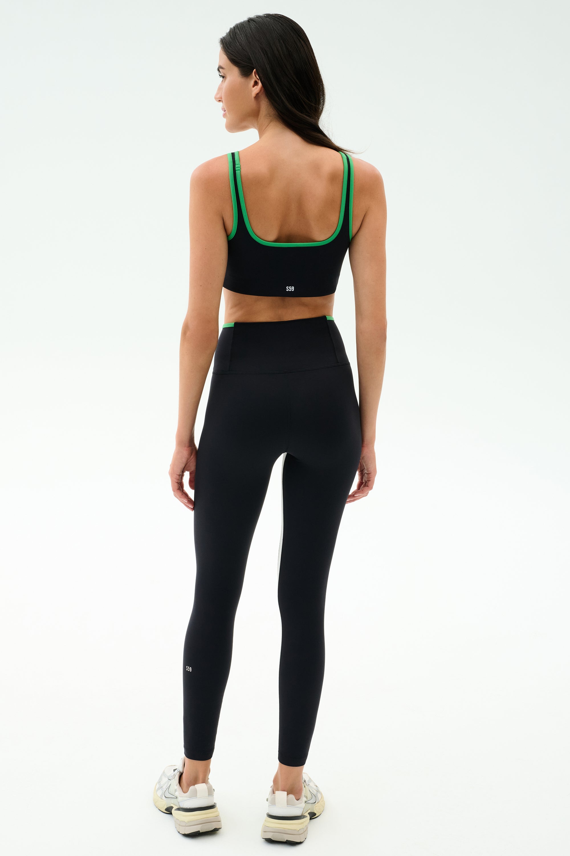 A woman faces away, wearing the SPLITS59 Cait Rigor Bra in Black/Grass, made from high-performance Rigor fabric, paired with high-waisted black leggings and white sneakers against a plain white background.