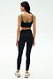 A woman faces away, wearing the SPLITS59 Cait Rigor Bra in Black/Grass, made from high-performance Rigor fabric, paired with high-waisted black leggings and white sneakers against a plain white background.