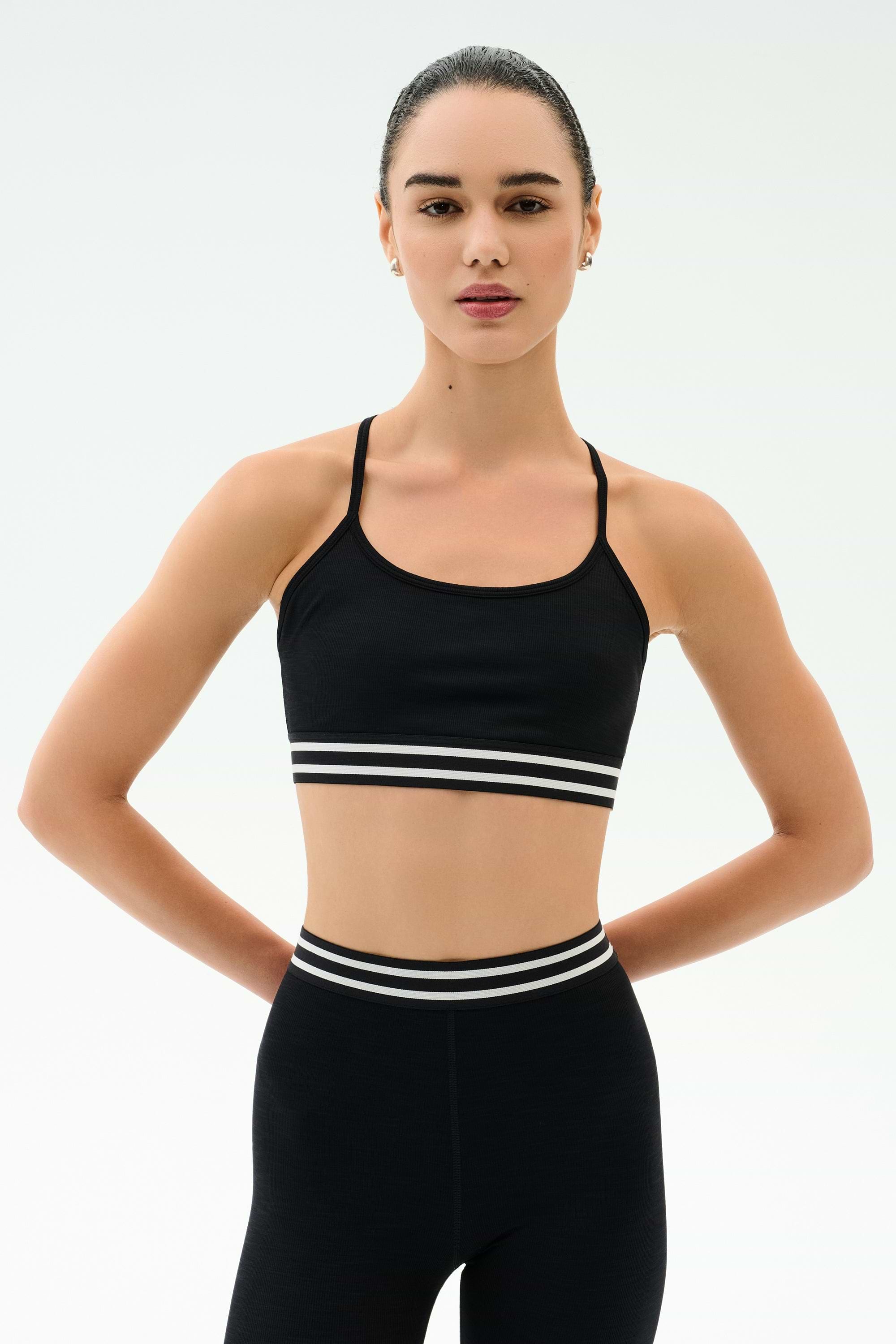 A woman with dark hair tied back wears the SPLITS59 Cali Active Rib Bra in black and matching high-waisted leggings, standing against a plain white background with her hands behind her back.