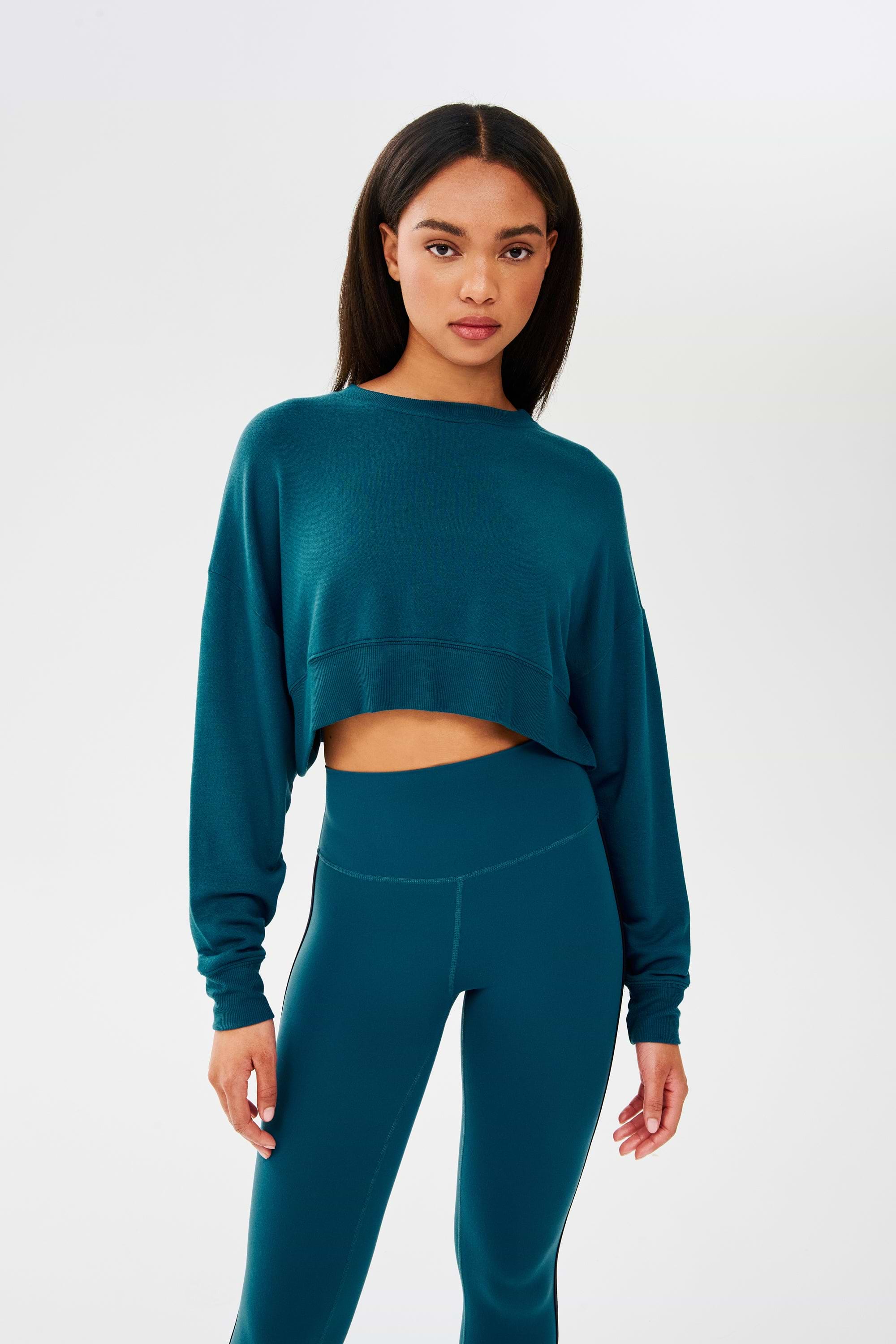 A woman wearing a SPLITS59 Noah Fleece Crop Sweatshirt in Peacock and leggings.