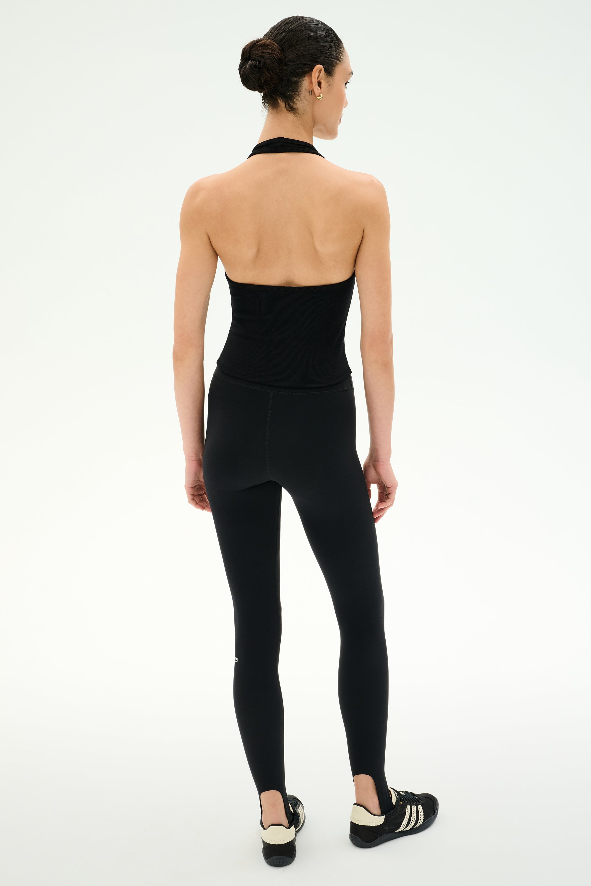 A person stands with their back to the camera, wearing the SPLITS59 June Rib Halter in black along with matching black leggings made from a soft modal blend, paired with black athletic shoes against a plain light background.