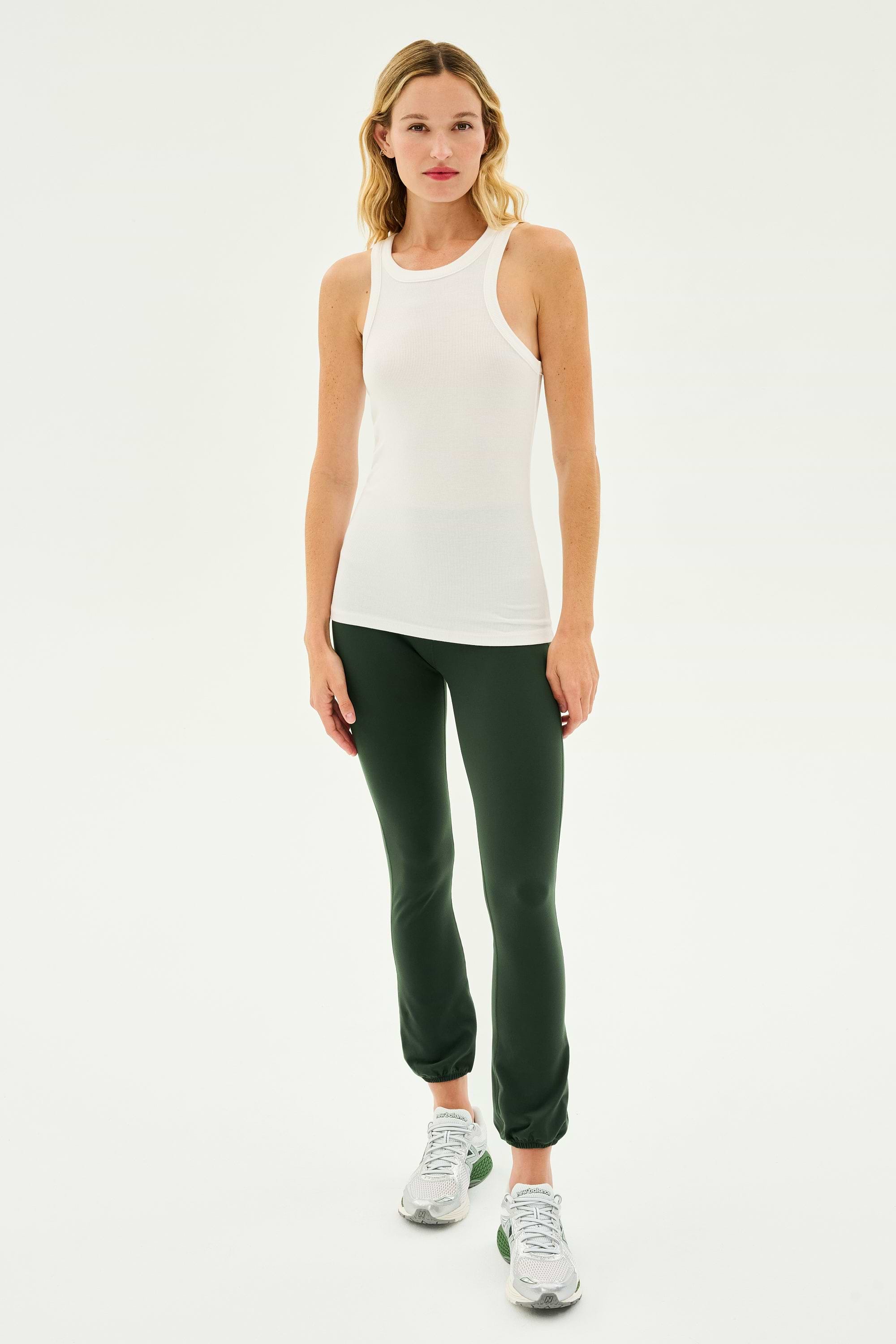 A woman stands facing forward wearing the Kiki Rib Tank Full Length in White by SPLITS59, dark green leggings, and gray athletic shoes against a plain white background, ready for yoga or gym workouts.