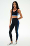 A woman is standing against a plain white background, wearing the SPLITS59 Ella High Waist Airweight 7/8 leggings in Black/Classic Blue with a matching black sports bra and white sneakers.