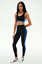 A person stands against a plain white background wearing the SPLITS59 Maci Airweight Bra in Black/Classic Blue, paired with matching nylon-spandex leggings featuring blue stripes and white sneakers.