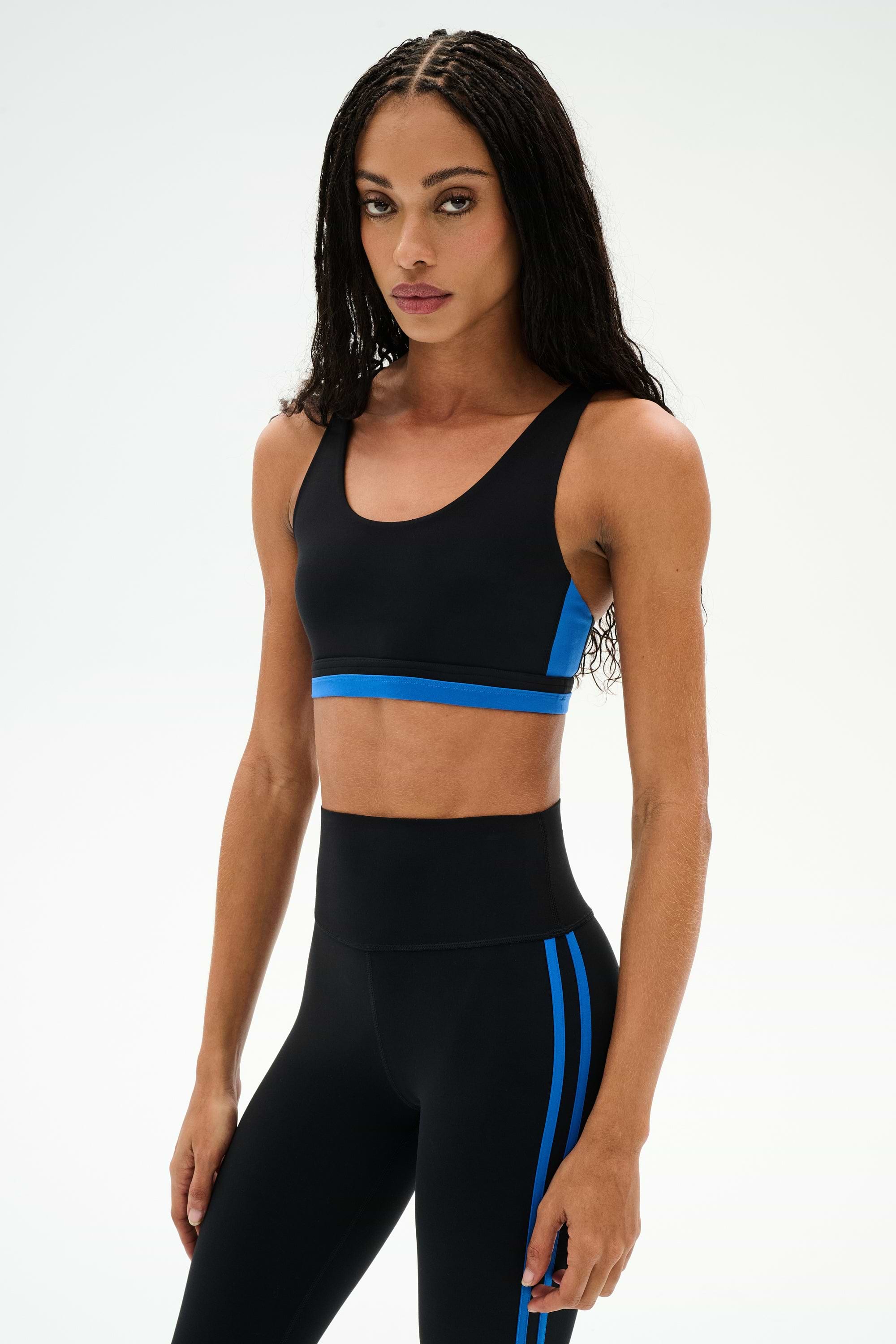 A woman wearing the SPLITS59 Maci Airweight Bra in Black/Classic Blue and coordinating nylon leggings with blue stripes, standing against a plain white background.