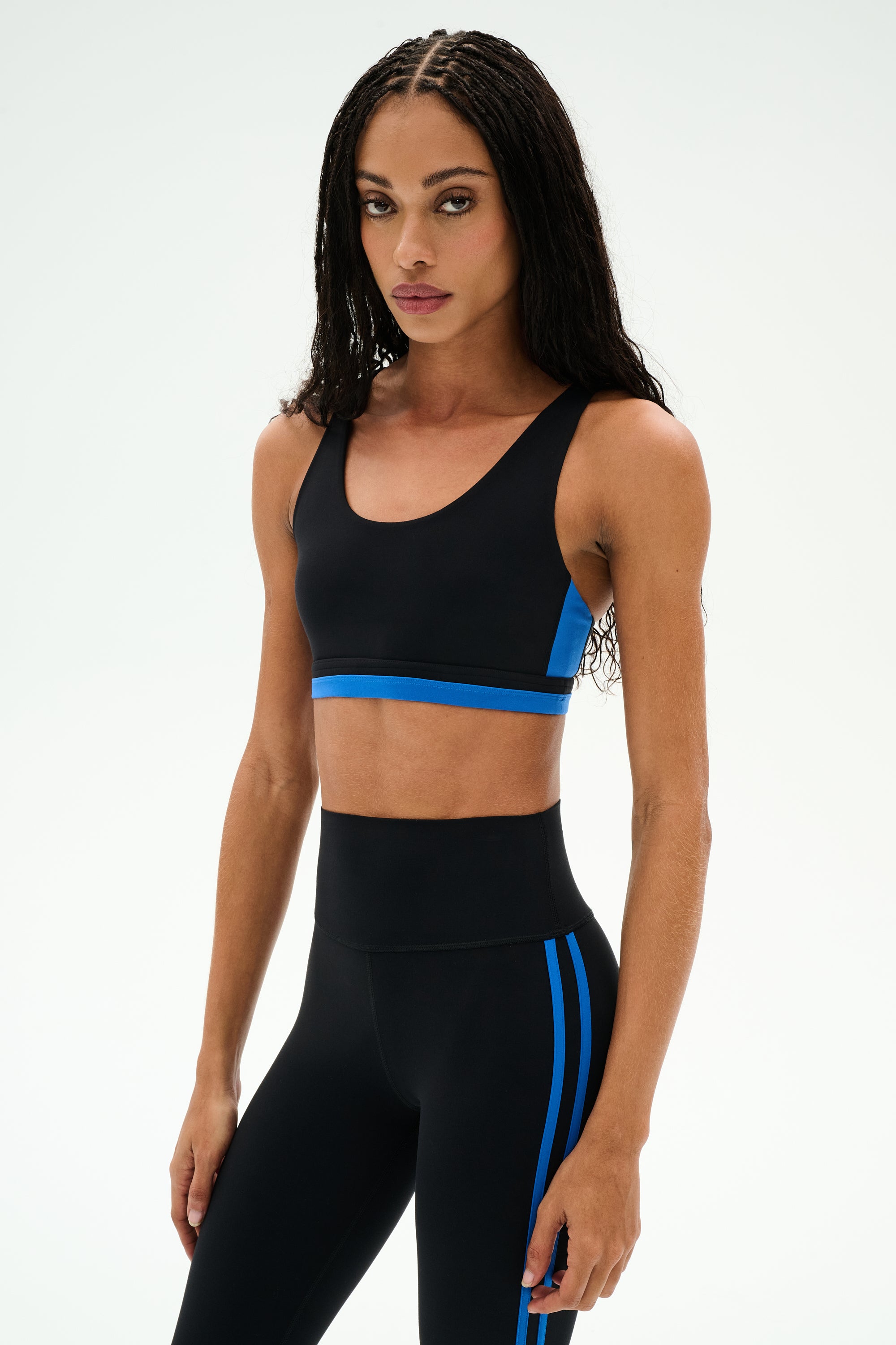 A woman wearing the SPLITS59 Maci Airweight Bra in Black/Classic Blue and coordinating nylon leggings with blue stripes, standing against a plain white background.