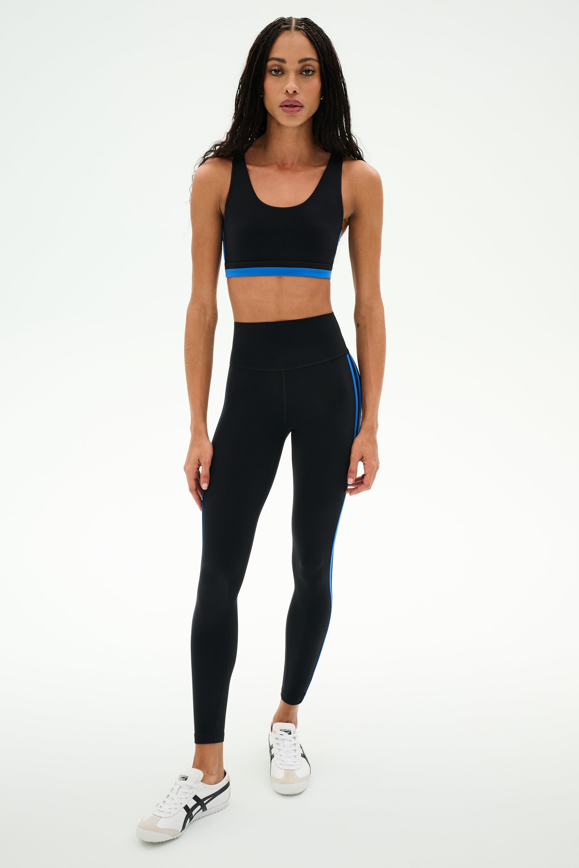 A woman wearing the SPLITS59 Maci Airweight Bra in Black/Classic Blue with matching black leggings featuring blue stripes, paired with white sneakers, stands against a plain white background.