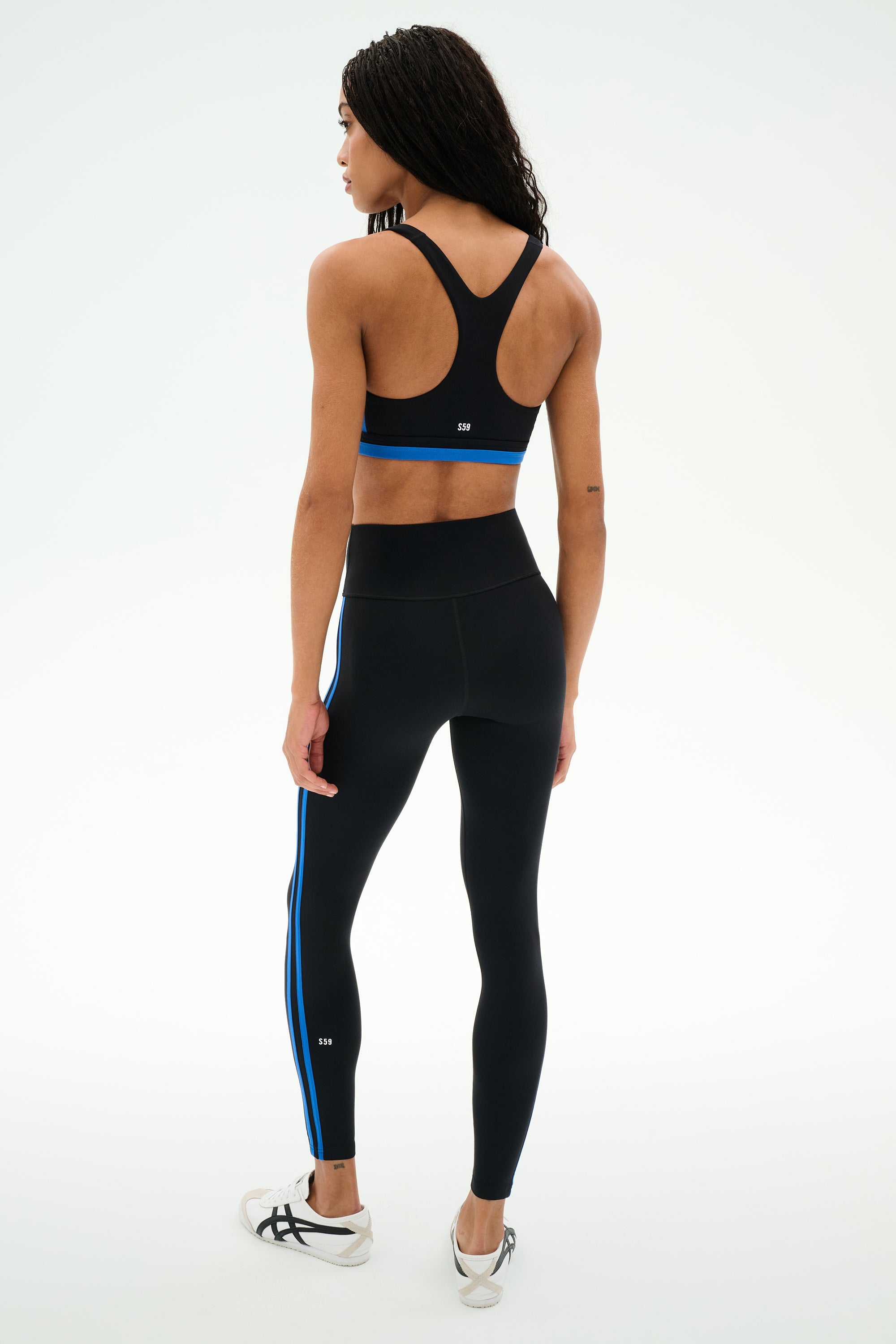 A person stands facing away against a plain white background, wearing the SPLITS59 Maci Airweight Bra in Black/Classic Blue.