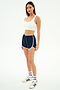 A woman stands against a white background wearing a white spandex crop top, SPLITS59 Megan Woven Nylon Run Short in Indigo/Creme, crew socks, and white Adidas sneakers.