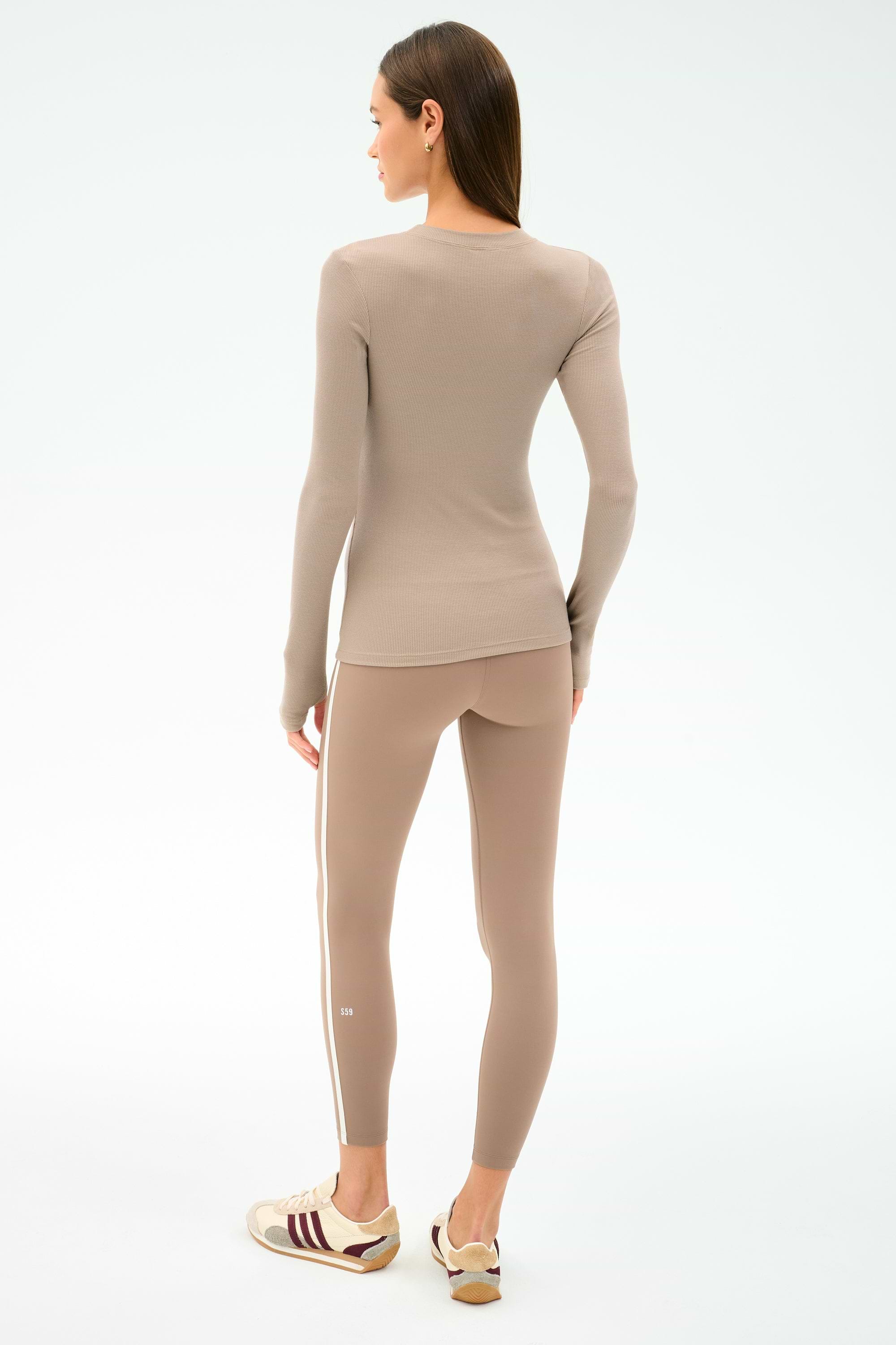 A woman faces away, wearing SPLITS59’s Louise Rib Long Sleeve in Latte and matching leggings with a white stripe—perfect for the gym—paired with beige sneakers with maroon stripes, on a plain light background. Made in Los Angeles.