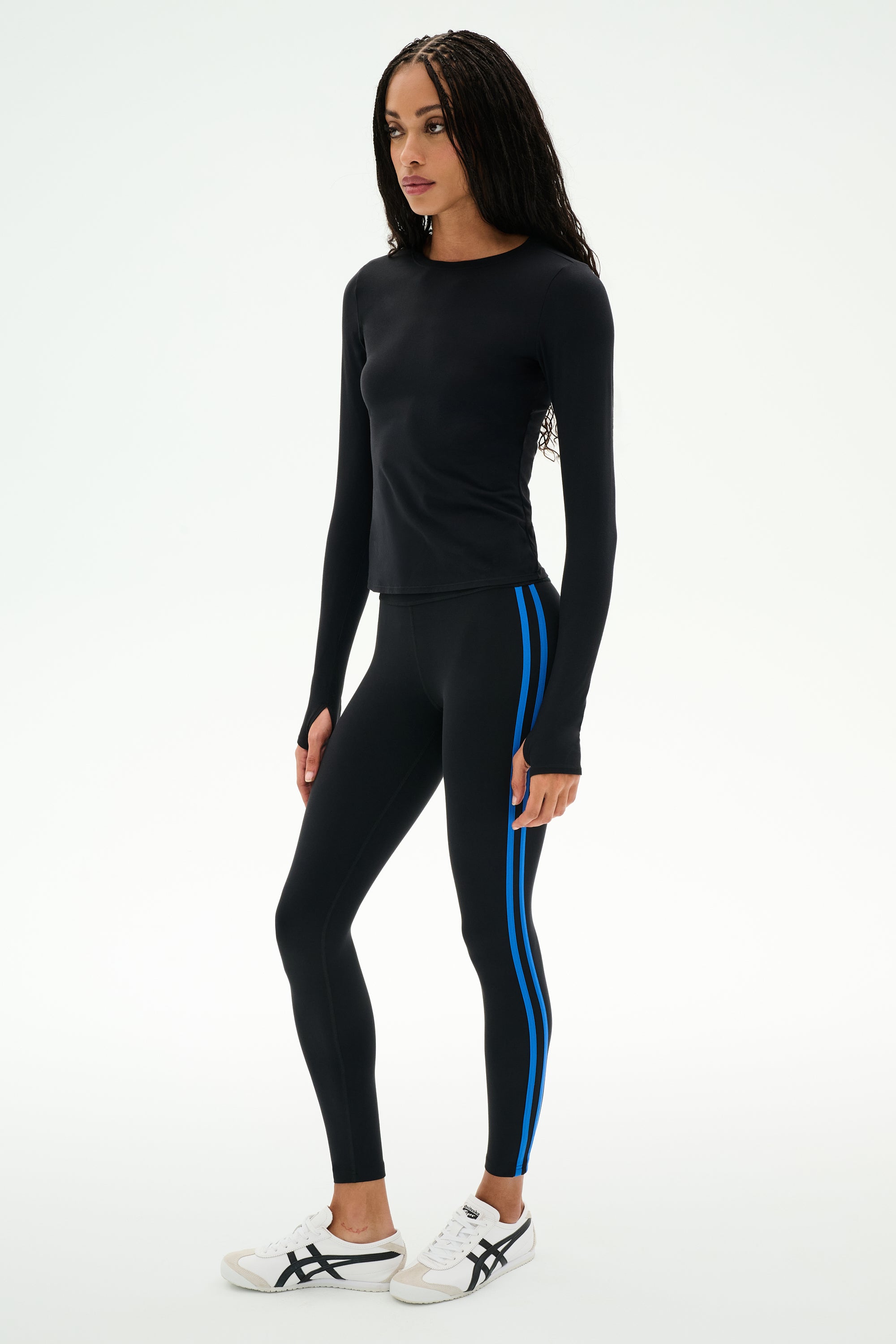 A woman is standing against a plain white background, dressed in the SPLITS59 Airweight Lite Long Sleeve Waist Length top in black, paired with black leggings featuring blue side stripes and white sneakers—an ideal outfit for gym workouts.