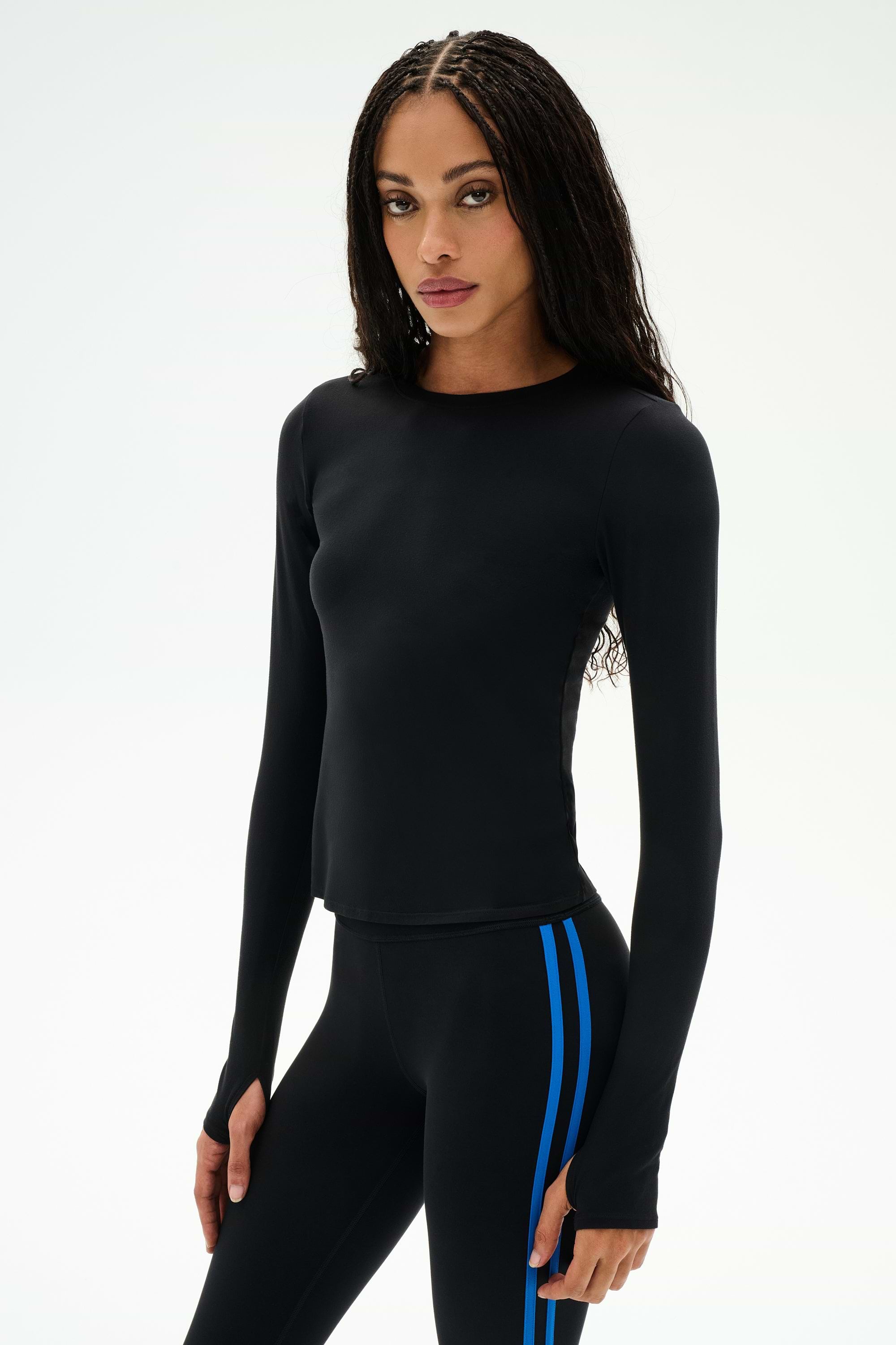 A woman with long braided hair models the SPLITS59 Airweight Lite Long Sleeve Waist Length top in black, paired with black leggings featuring blue side stripes. The fitted, quick-dry, and lightweight workout tee is perfect for gym sessions. She stands confidently against a plain white background.