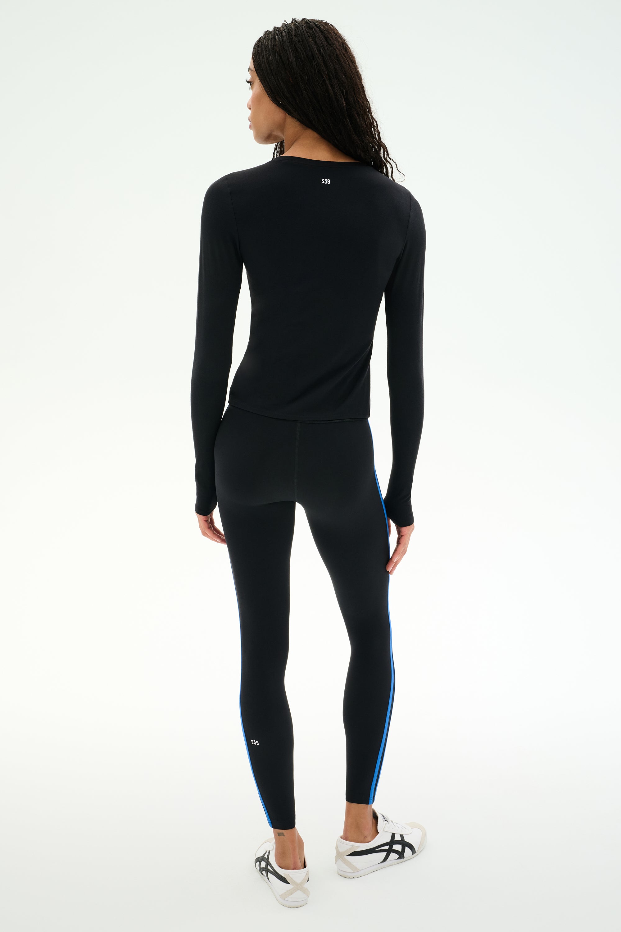 A person stands facing away, wearing the SPLITS59 Airweight Lite Long Sleeve Waist Length top in black and matching leggings with blue side stripes, paired with white sneakers against a plain light background—an ideal outfit for gym workouts.