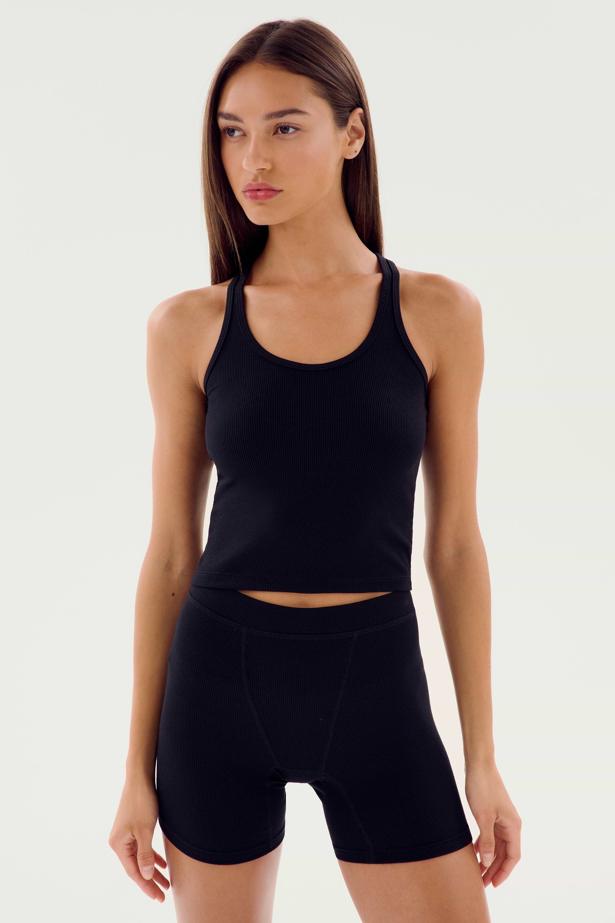 Woman wearing the SPLITS59 Ashby Rib Crop, a fitted black tank top, paired with black high-waisted shorts, standing against a plain light background.