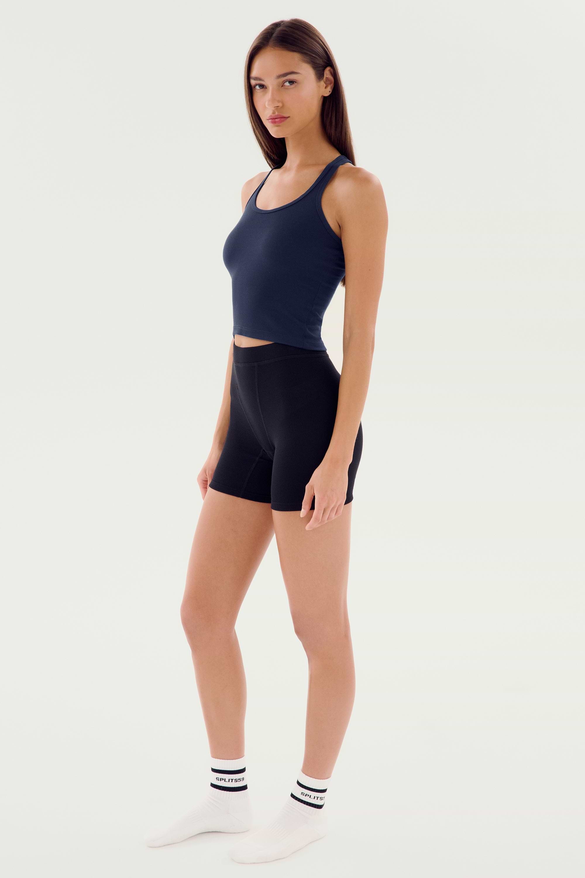 A person stands against a plain background, wearing a navy Ashby Rib Crop - Indigo by SPLITS59, black biker shorts, white socks, and white shoes. They are looking towards the camera with a neutral expression.