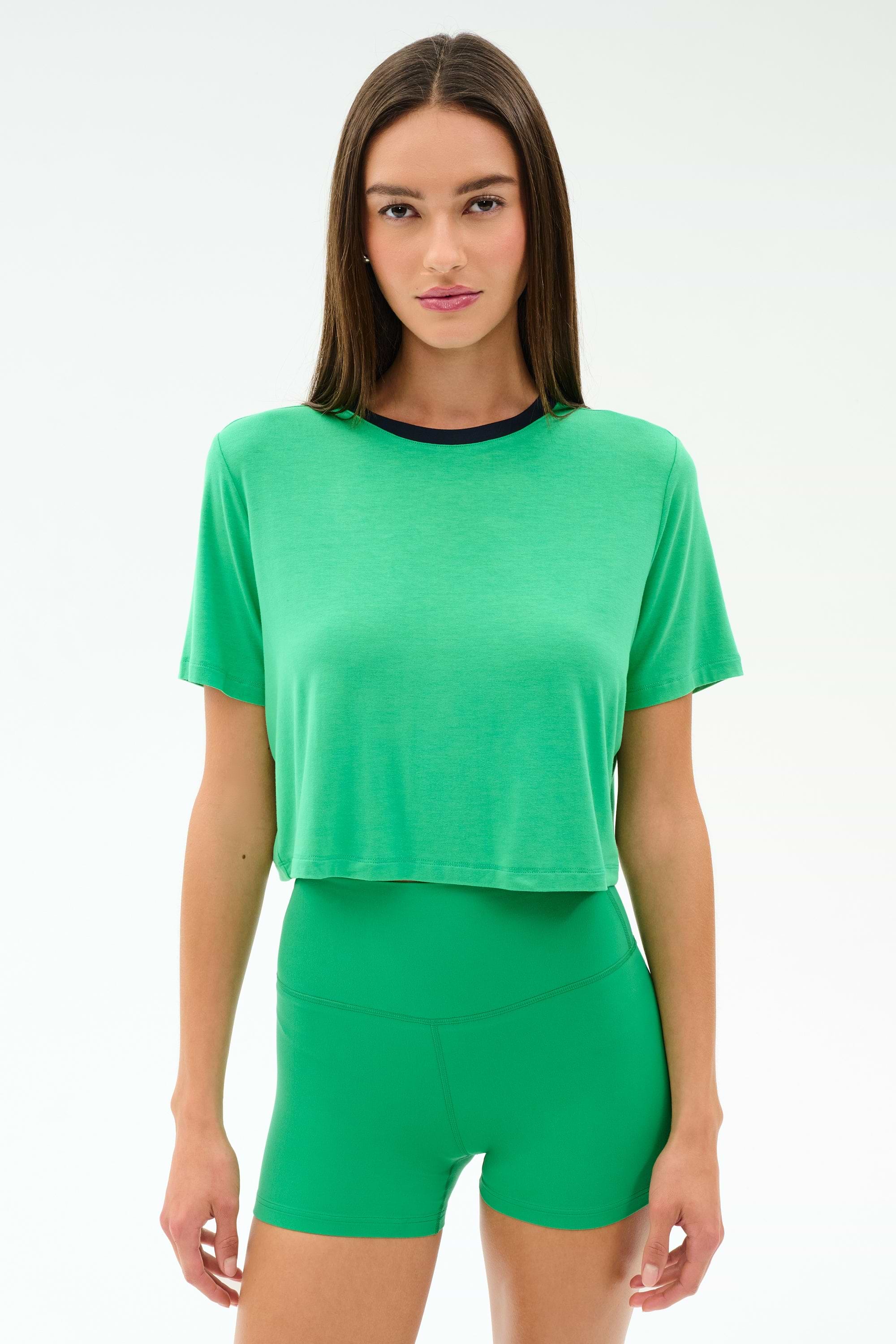 A woman wears the SPLITS59 Djuna Crop Ringer in Spearmint/Indigo, paired with green high-waisted shorts, both crafted from soft viscose fabric, standing against a plain light background.