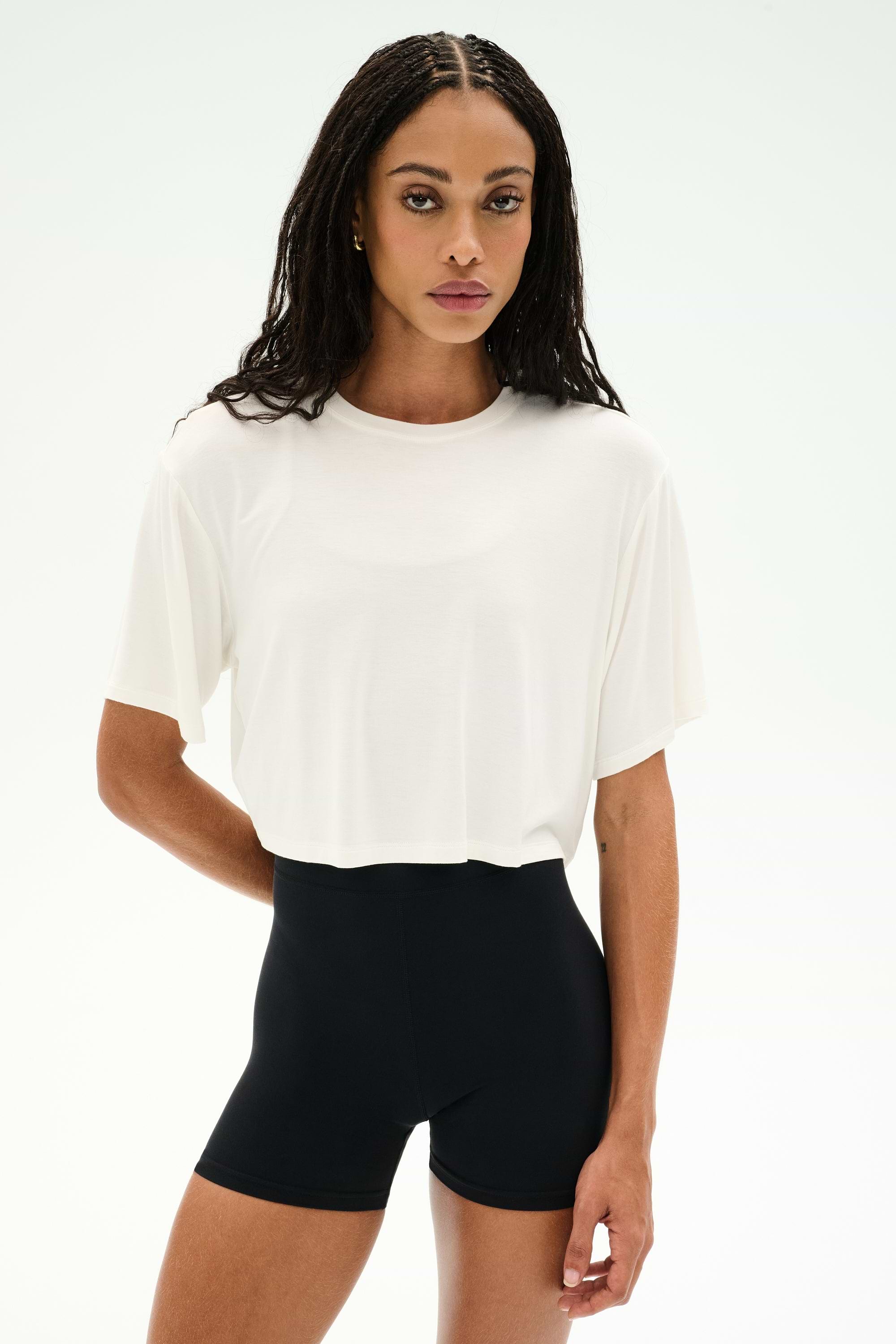 A woman faces forward against a white background, wearing the SPLITS59 Djuna Oversized Crop Jersey Tee in white paired with high-waisted black stretchy shorts.